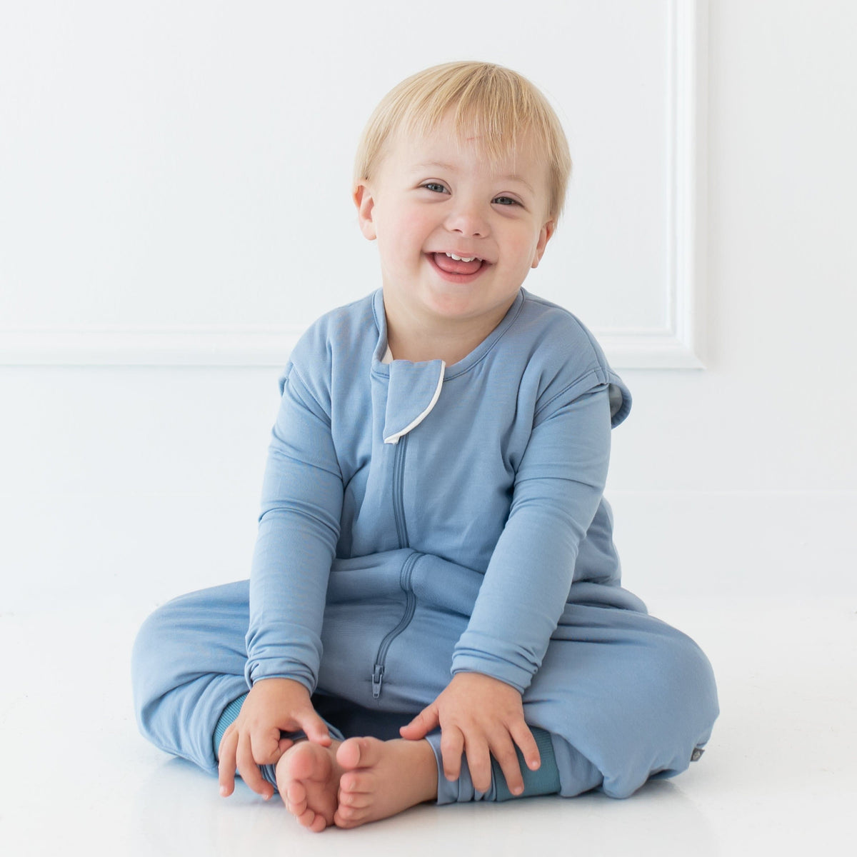 Smiling toddler sitting on floor and wearing the Sleep Bag Walker in Slate 1.0
