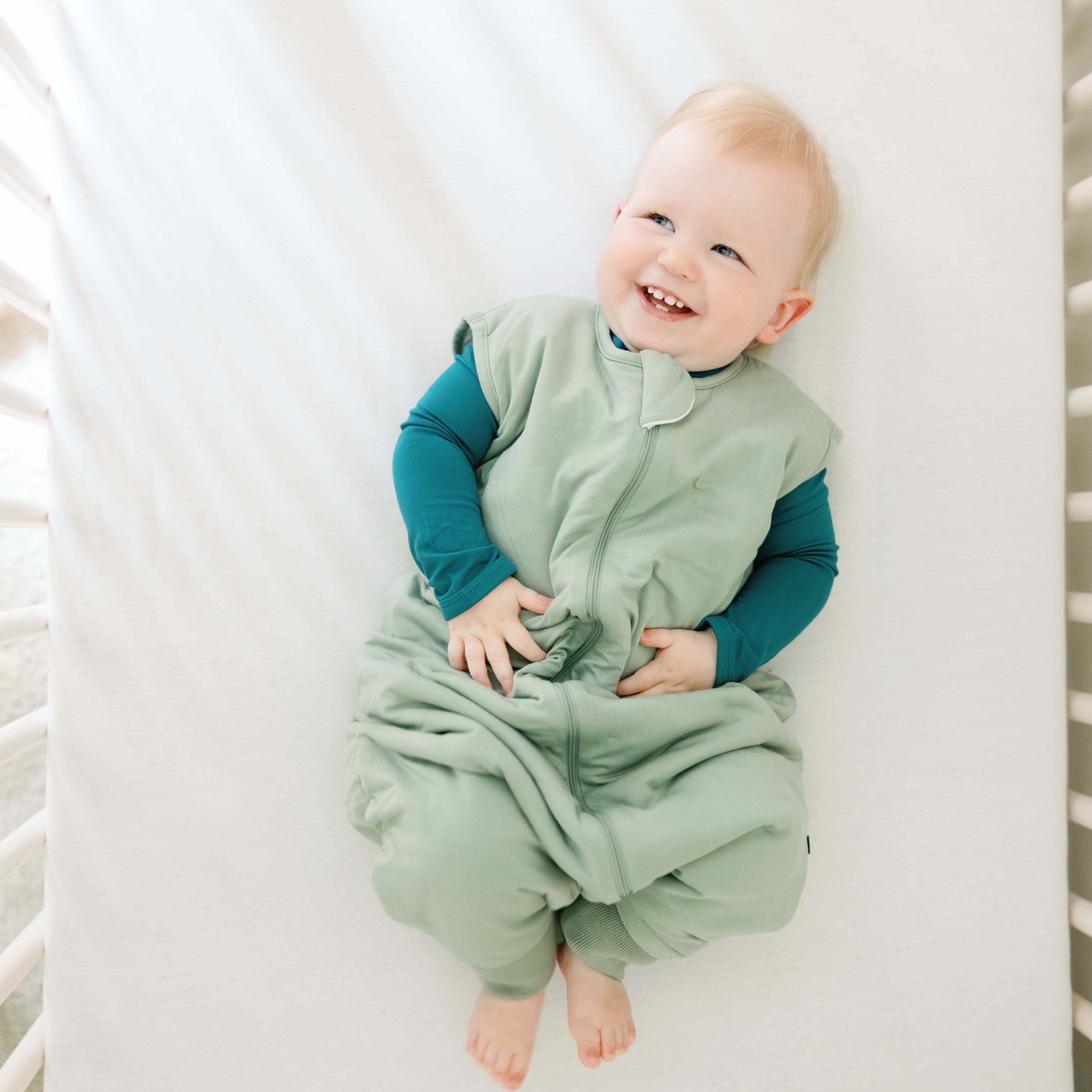 Smiling infant laying in a crib wearing the Sleep Bag Walker in Thyme 1.0