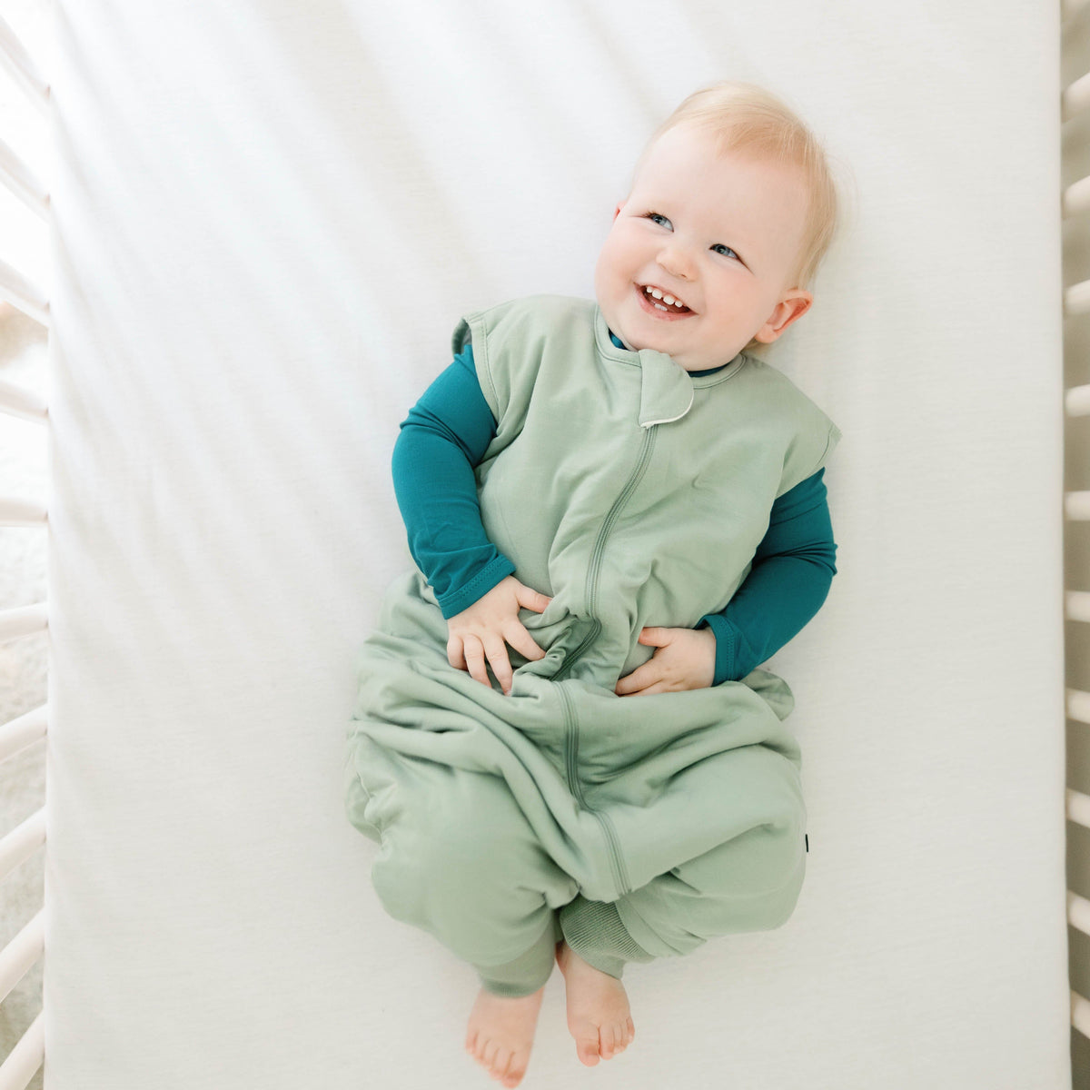 Smiling infant laying in a crib wearing the Sleep Bag Walker in Thyme 1.0