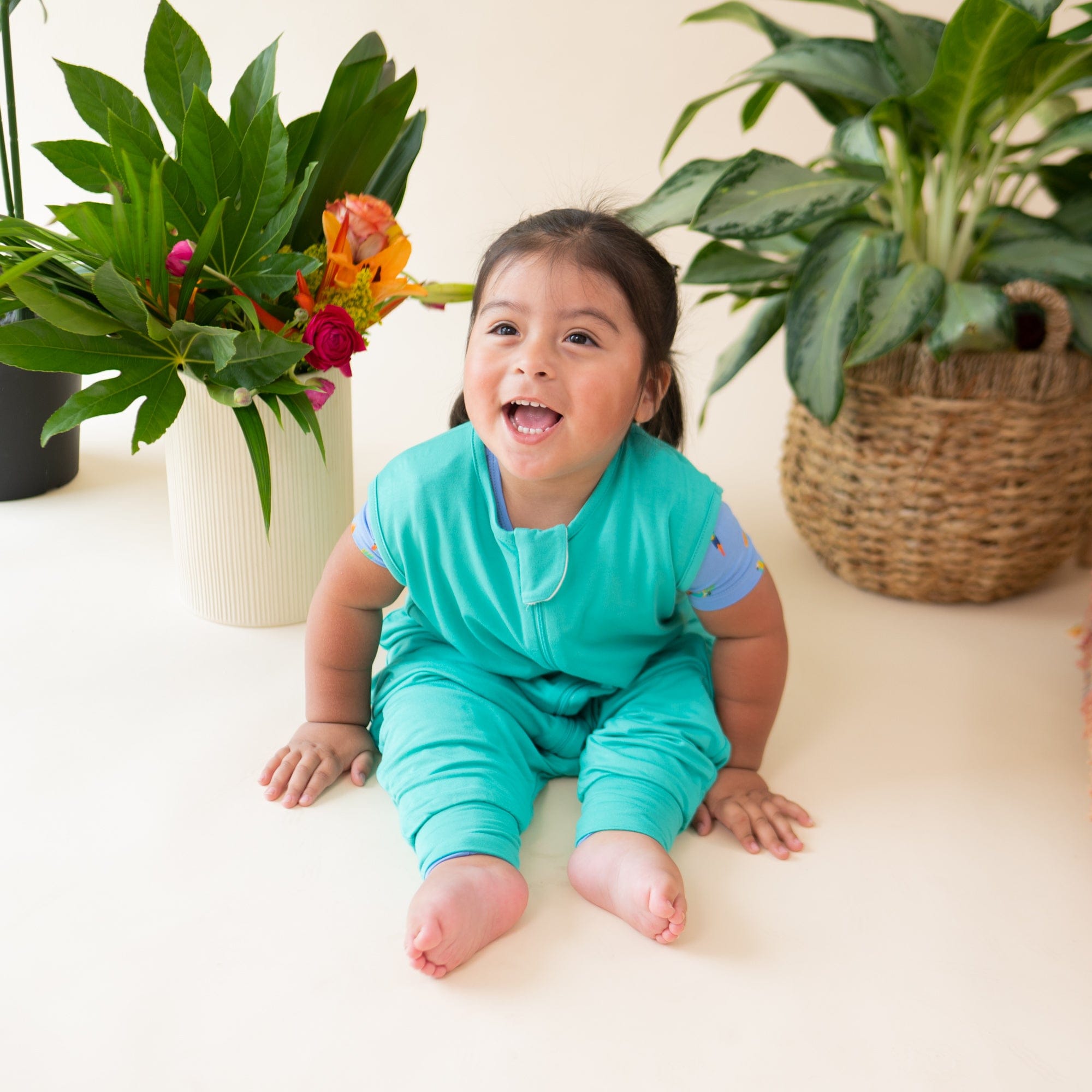 Smiling model sitting wearing the Sleep Bag Walker in Caribbean 0.5 in front of plotted plants