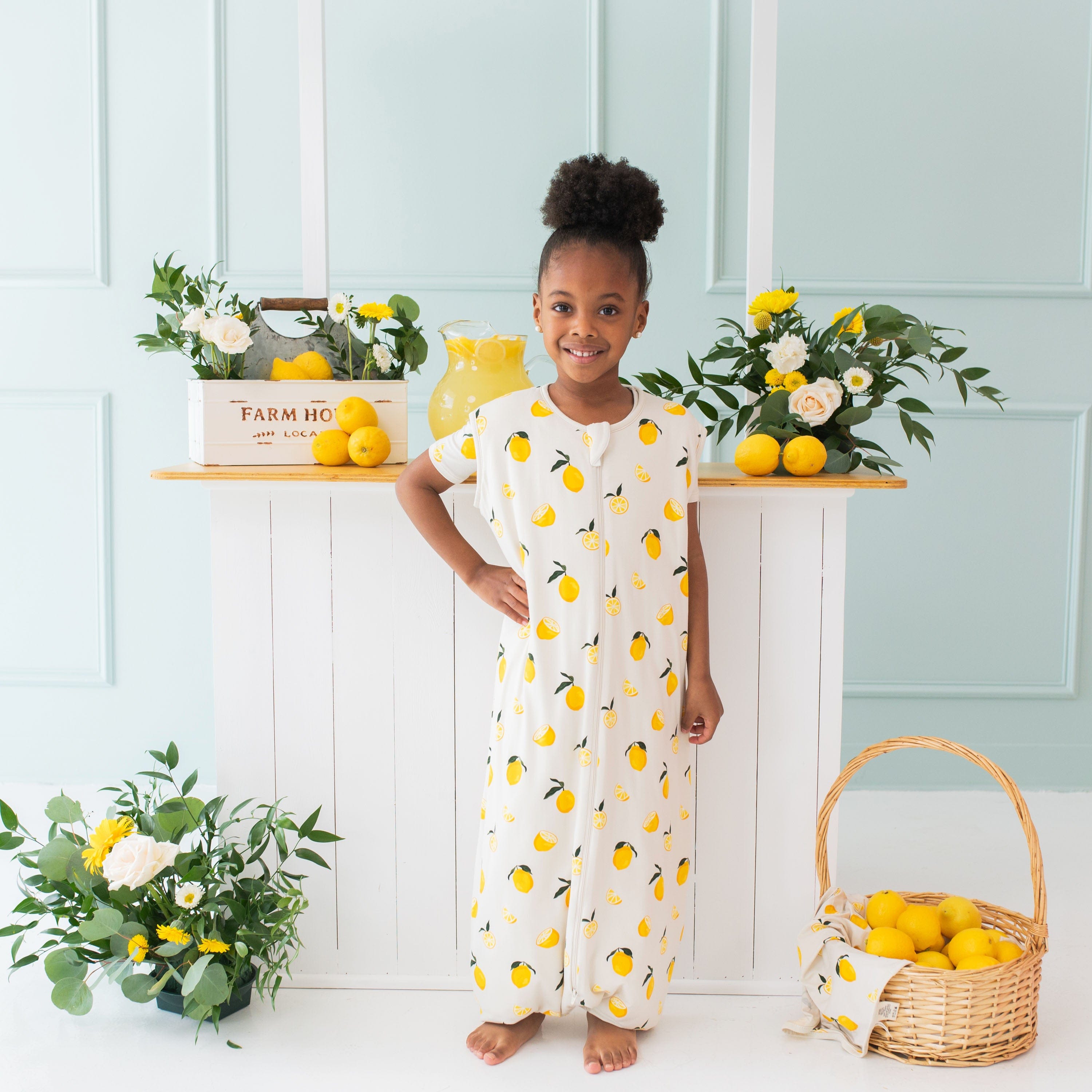 Young girl model standing in front of a wooden lemonade stand with one hand on her hip wearing the Sleep Bag Walker in Lemon 1.0