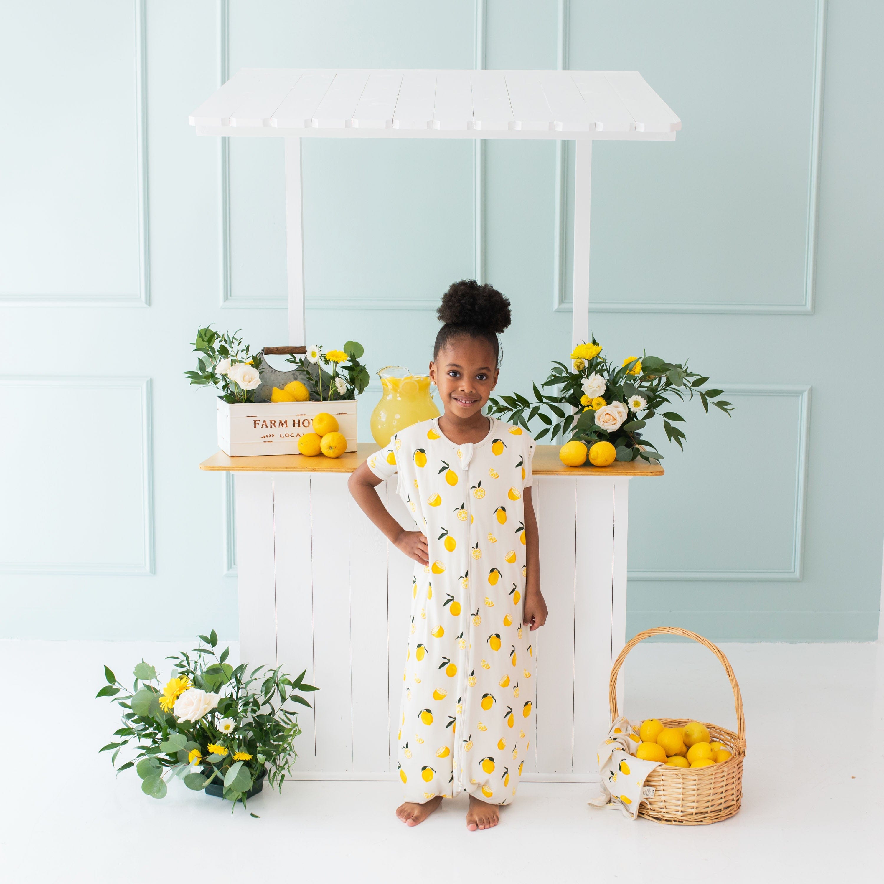 Young girl model standing with her hand on her hip in front of a lemonade stand wearing the Sleep Bag Walker in Lemon 1.0