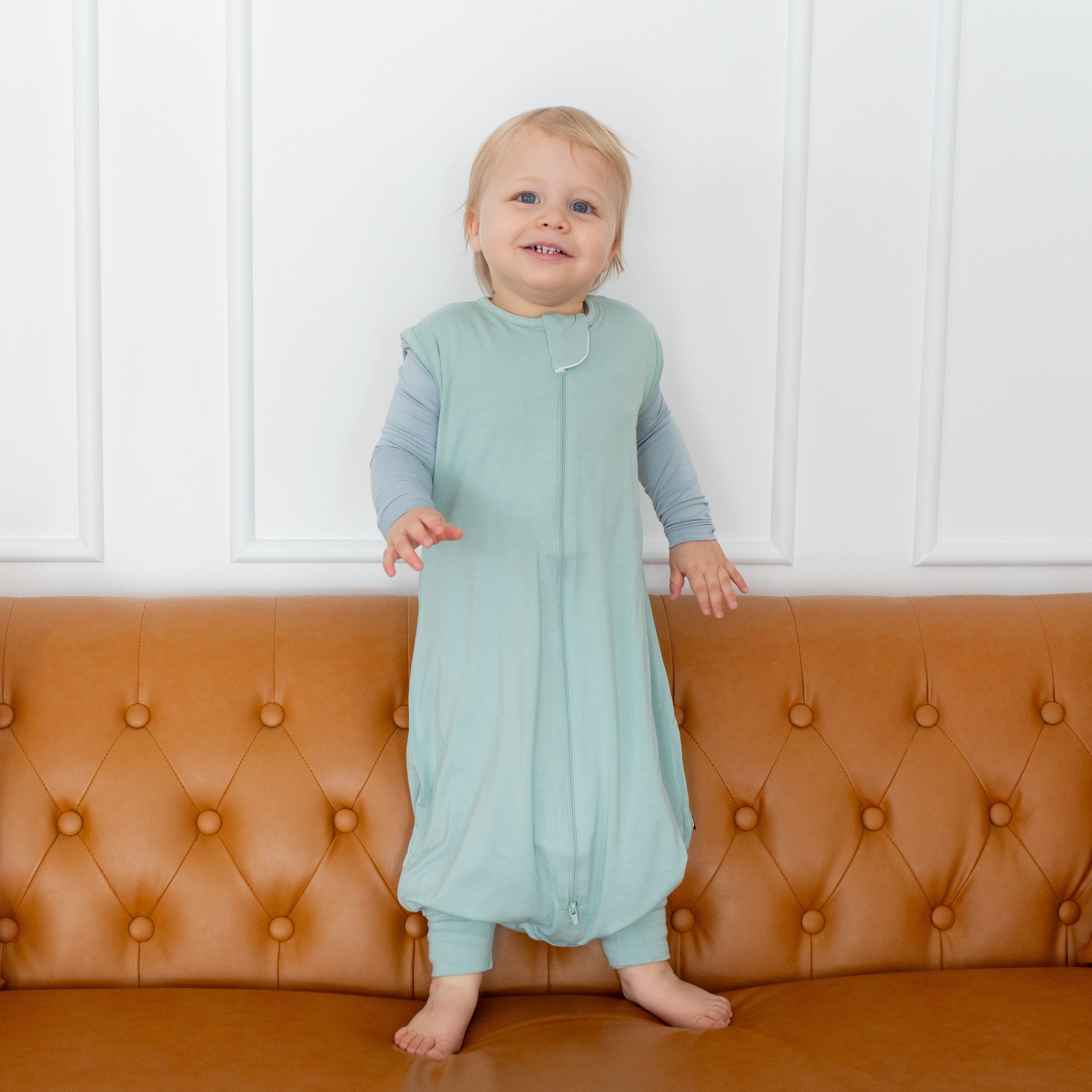 Toddler wearing Kyte Baby Sleep Bag Walker in Sage 0.5 while standing on a couch