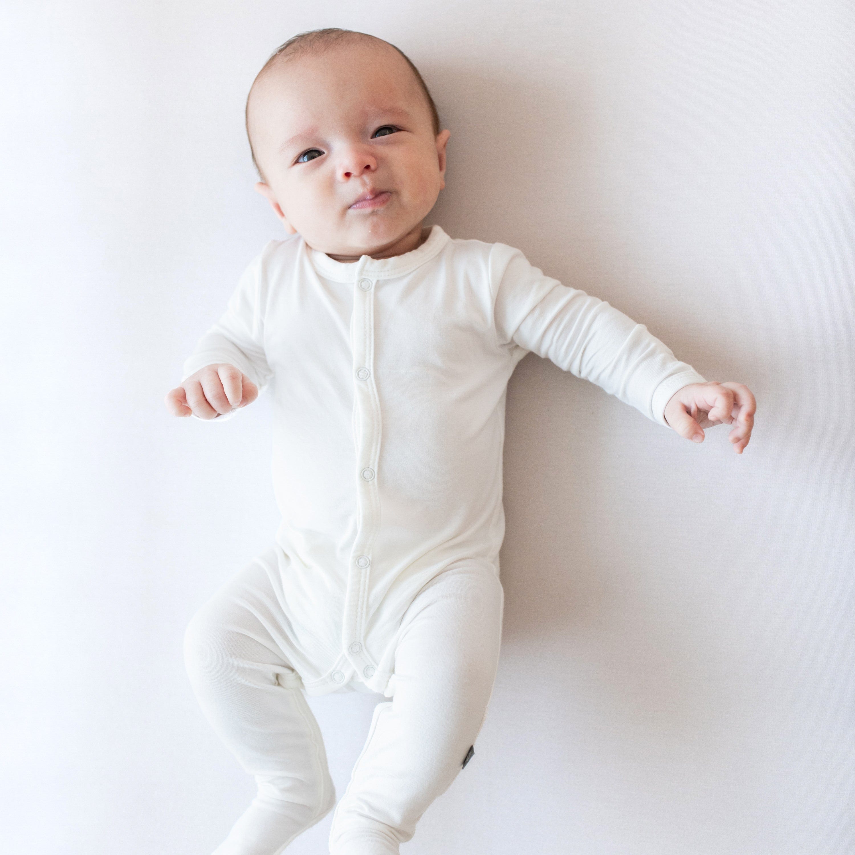 Baby wearing Kyte Baby Snap Footie in Cloud. Laying on white surface and looking at the camera