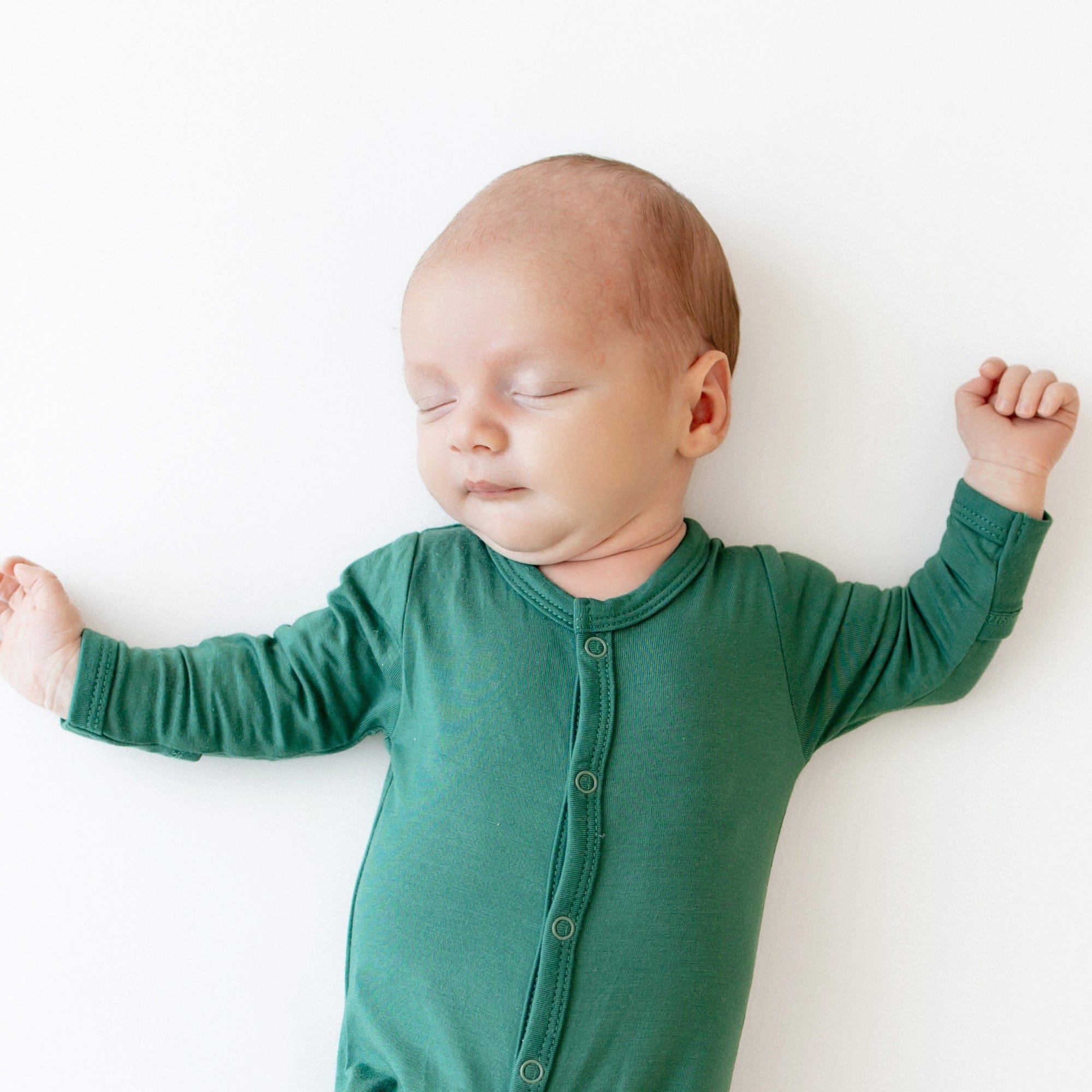 Close up of sleeping newborn laying on white surface while wearing Kyte Baby Footie in Emerald