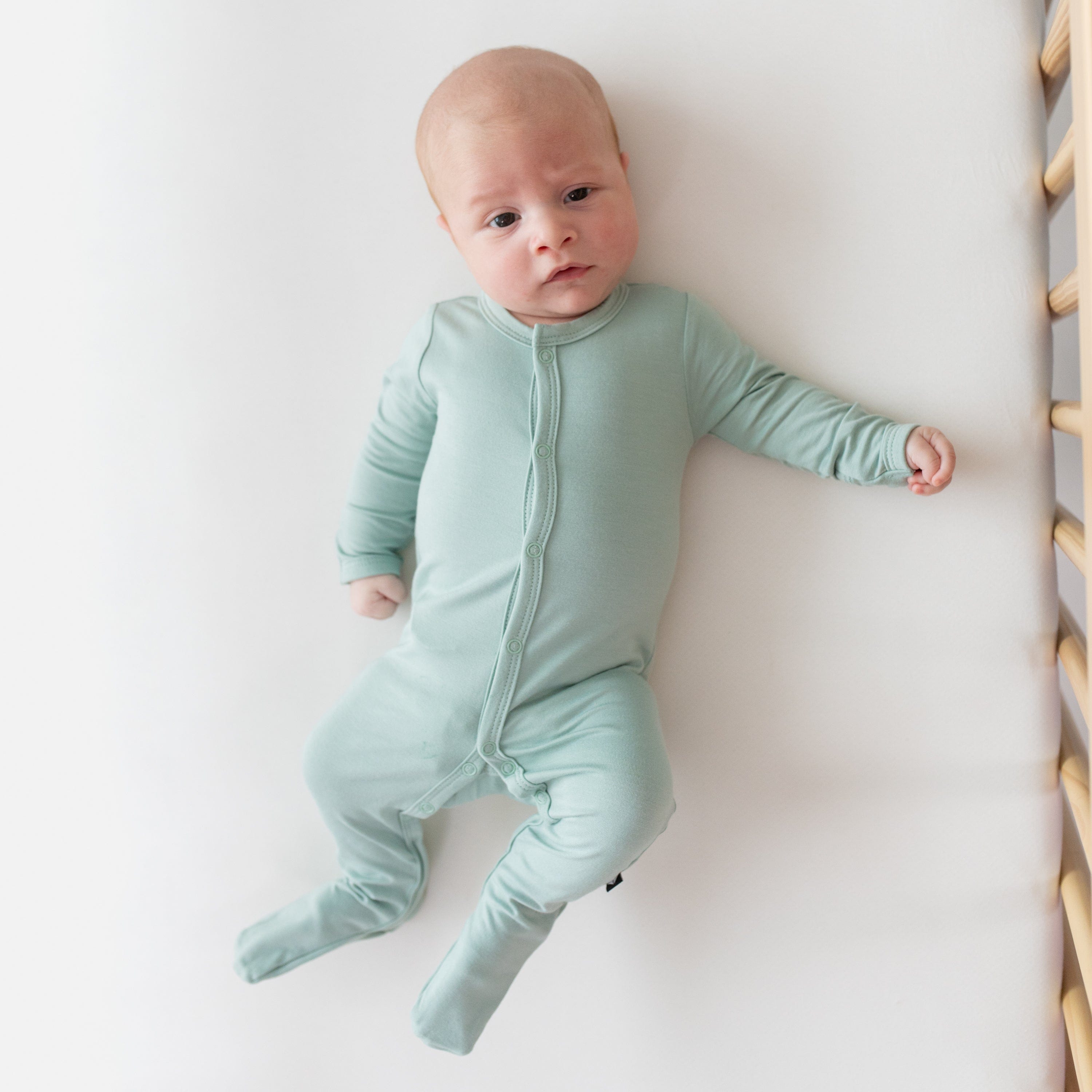 Newborn laying in crib and wearing Kyte Baby Footie in Sage