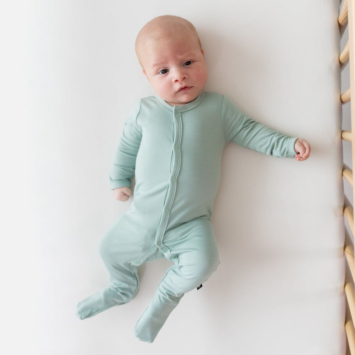 Newborn laying in crib and wearing Kyte Baby Footie in Sage