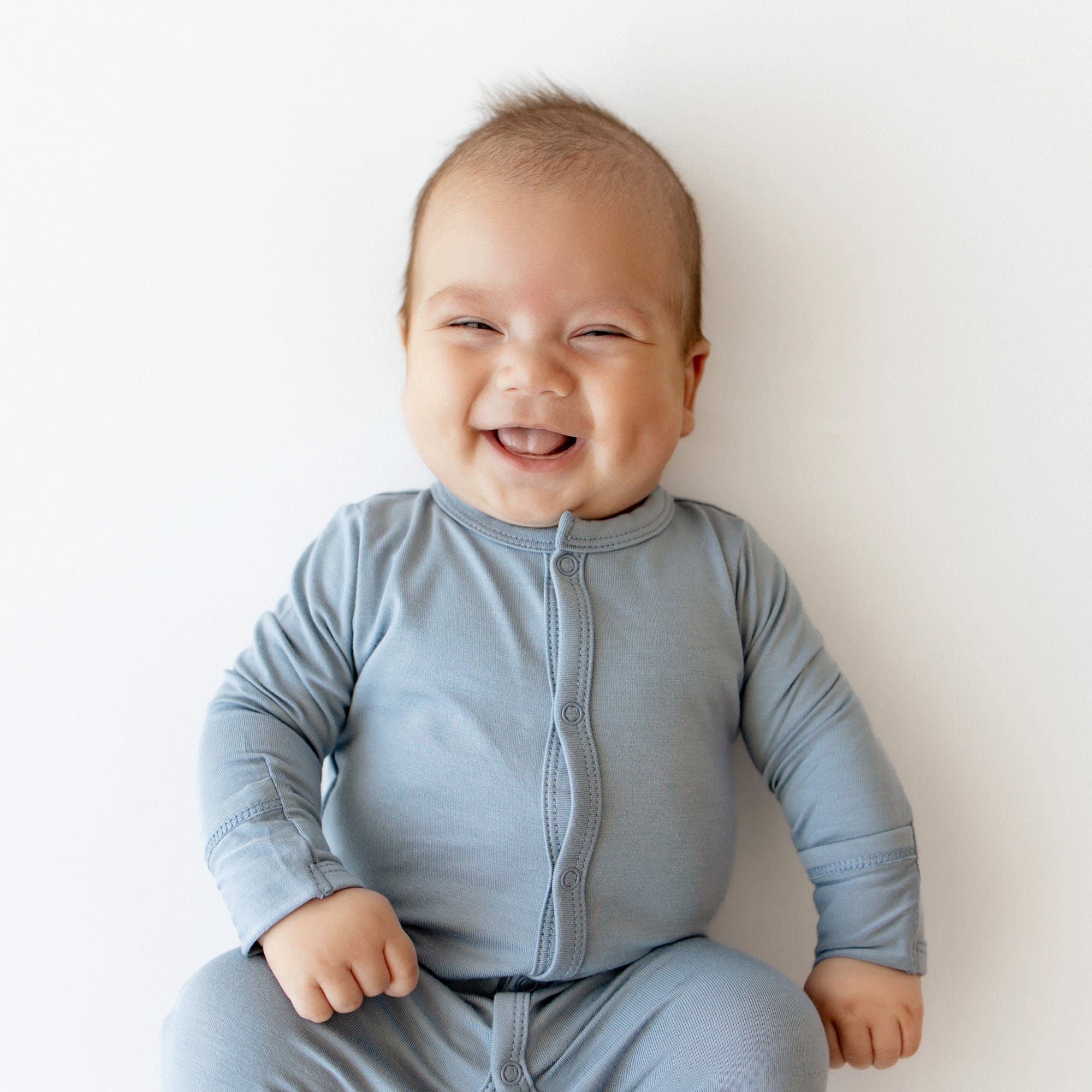 Close up of smiling baby wearing Kyte Baby Footie in Slate while laying on an off-white surface