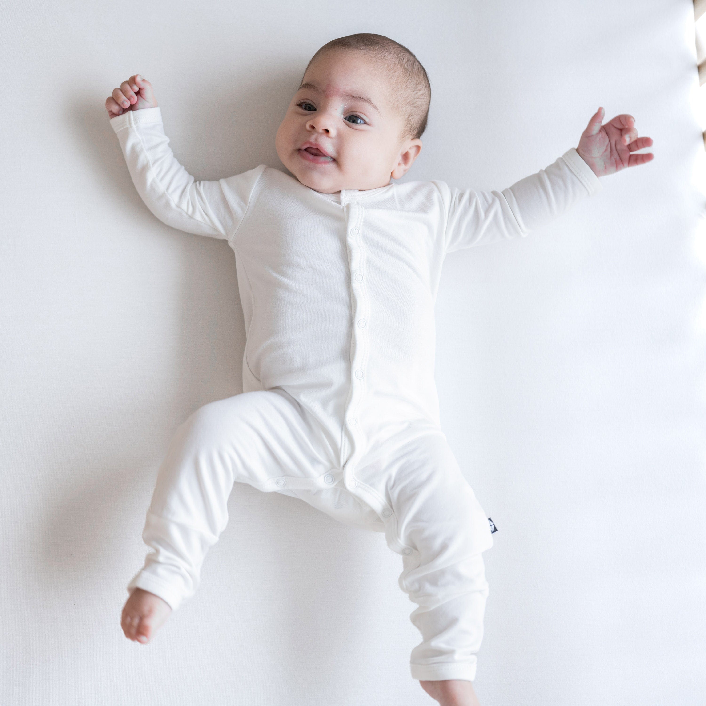 Infant lying on a white surface wearing Kyte Baby Snap Romper Romper in Cloud. Infant is smiling with arms and legs stretched out.
