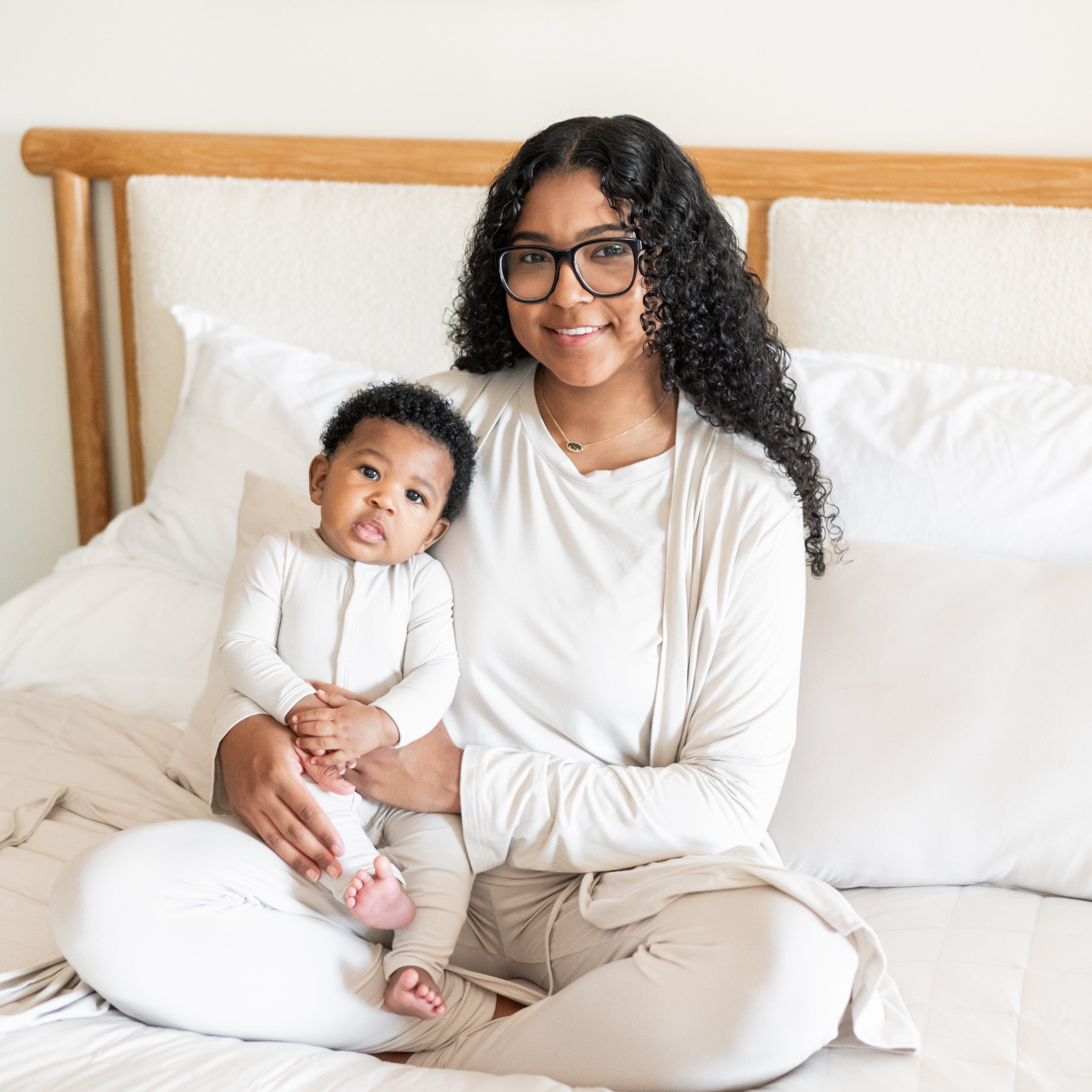 Mother and baby sitting on a bed matching while baby wears a Kyte Baby Snap Romper in Oat