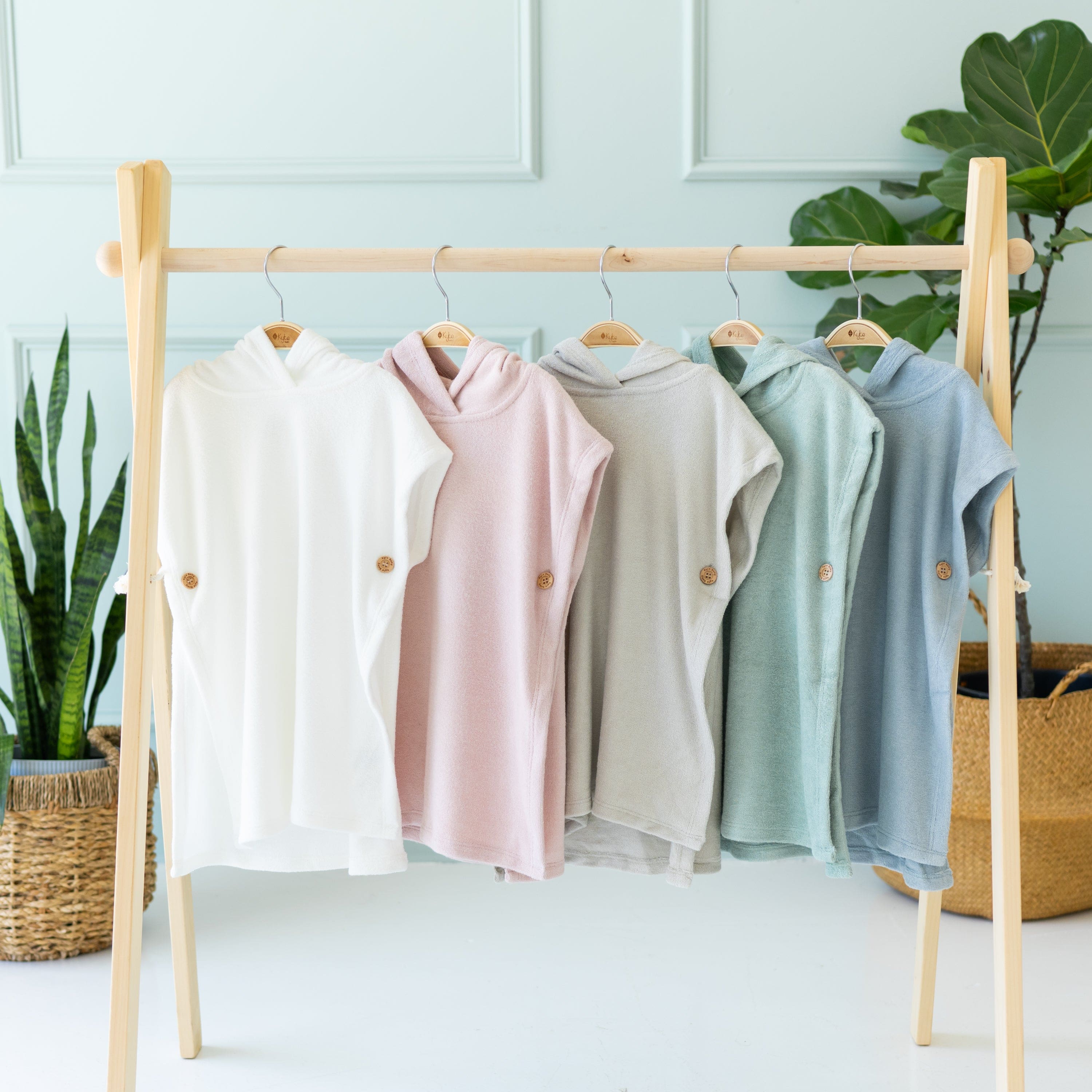 Multiple Kyte Baby Swim Ponchos in core colors hung on hangers on a wooden rack