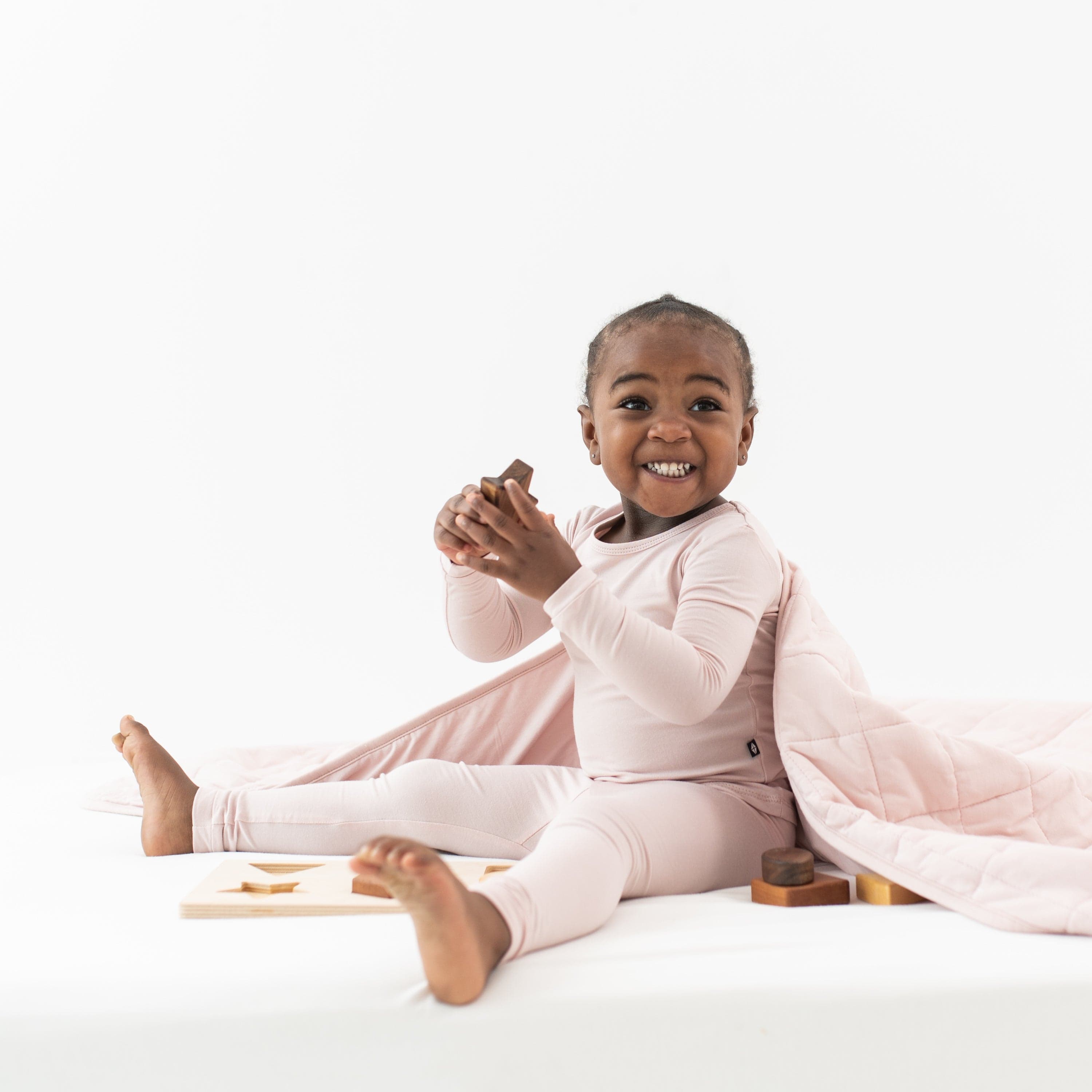 Smiling child playing with wooden toys and wrapped in a Toddler Blanket in Blush 1.0 TOG