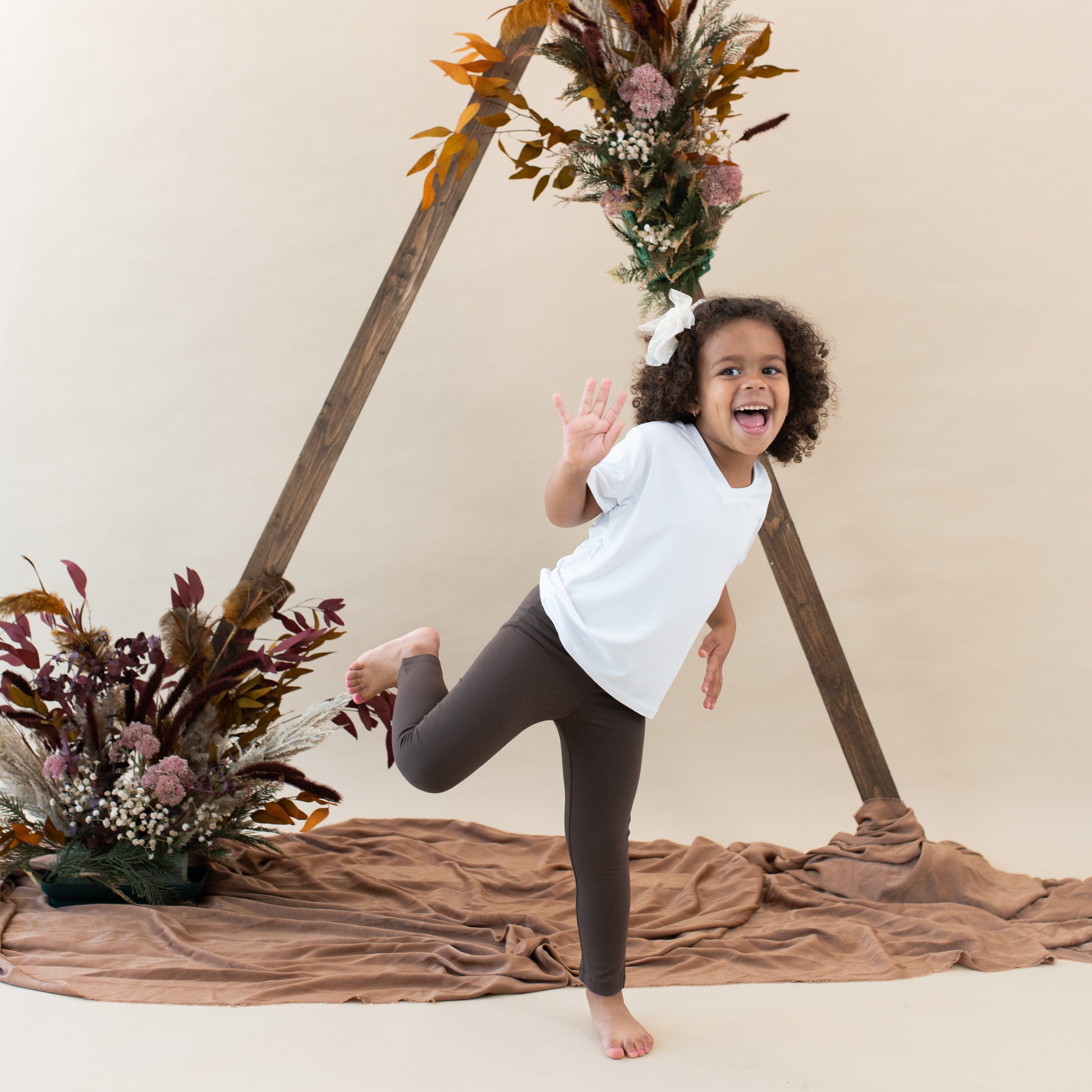 Girl model standing on one foot waving wearing the Toddler Leggings in Espresso paired with a Snow toddler V-neck