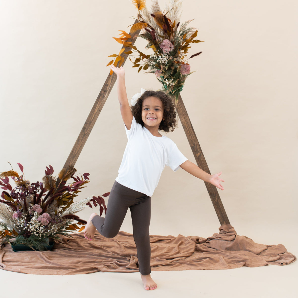 Girl model wearing the Toddler Leggings in Espresso paired with a Snow v-neck in front of a rustic flower backdrop