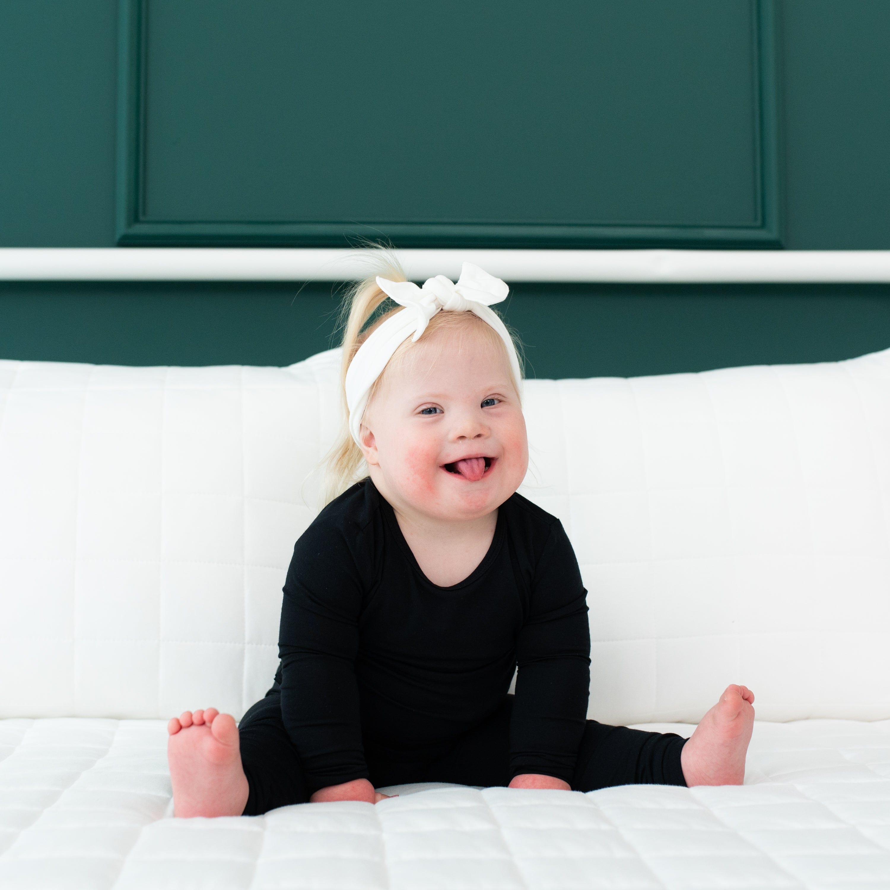 Toddler sitting on a bed while wearing a Long Sleeve Pajama Set in Midnight and Headband Bow in Cloud