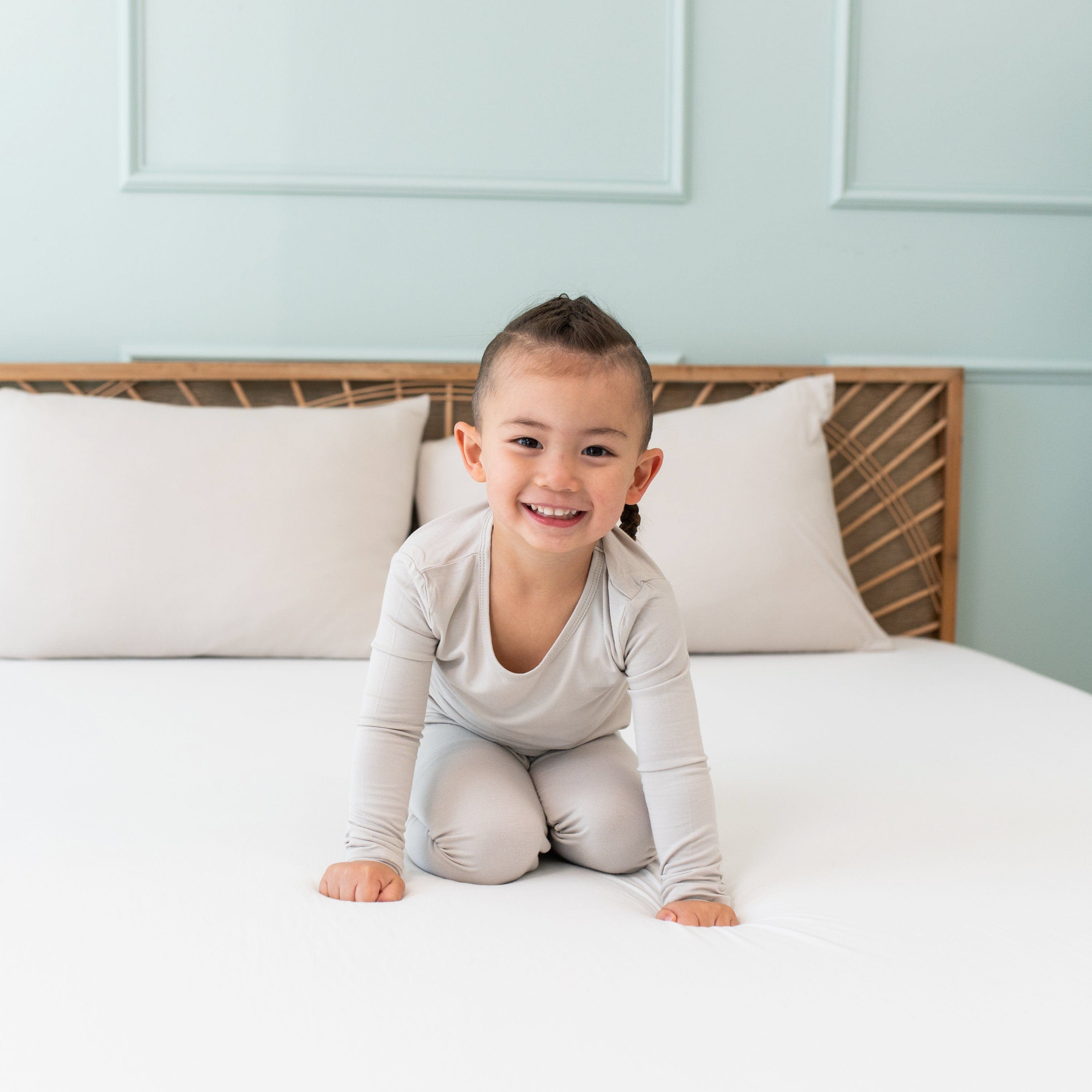 Toddler crawling on a bed with white sheets while wearing Kyte Baby Long Sleeve Pajamas in Oat