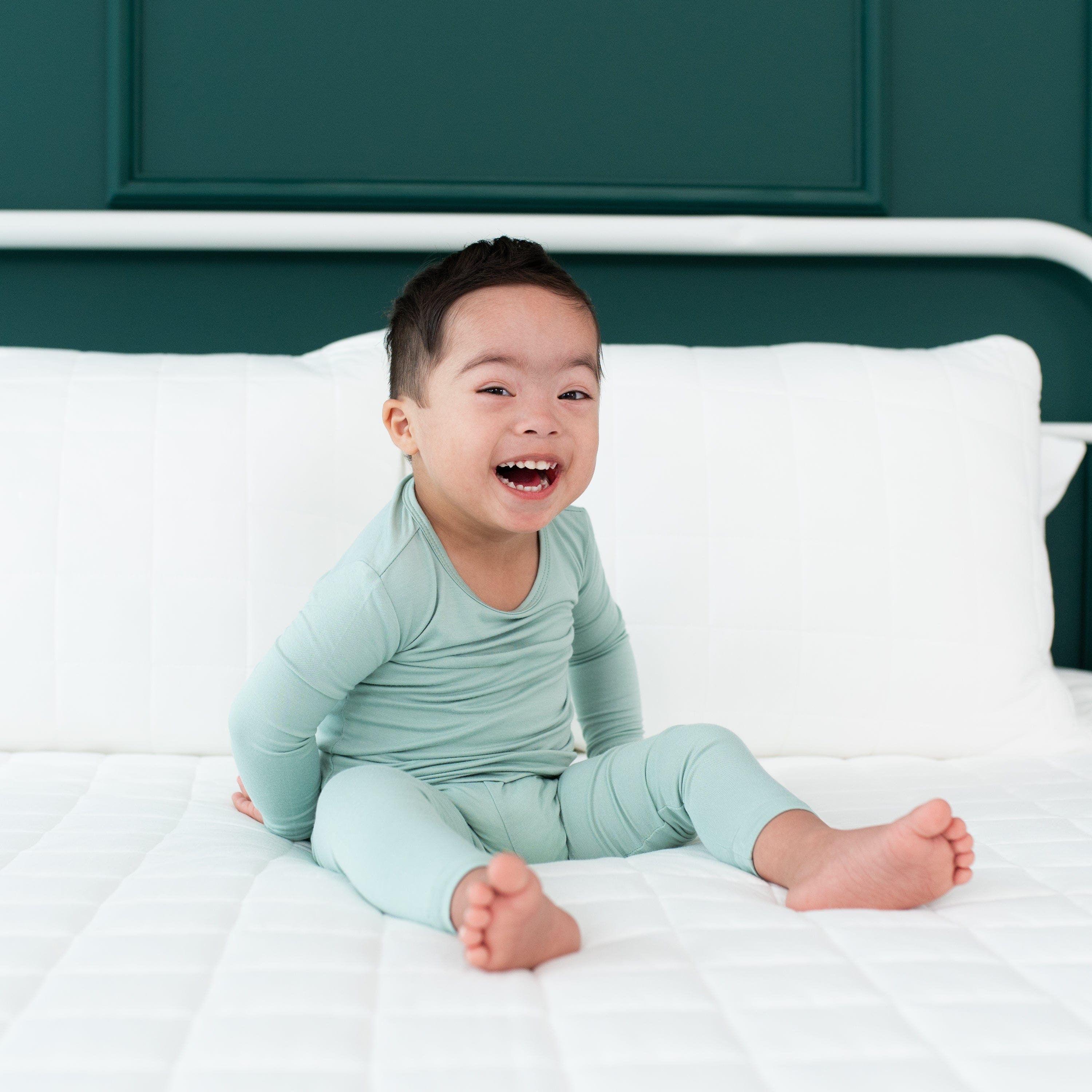 Smiling toddler sitting on a white bed while wearing Long Sleeve Pajama Set in Sage