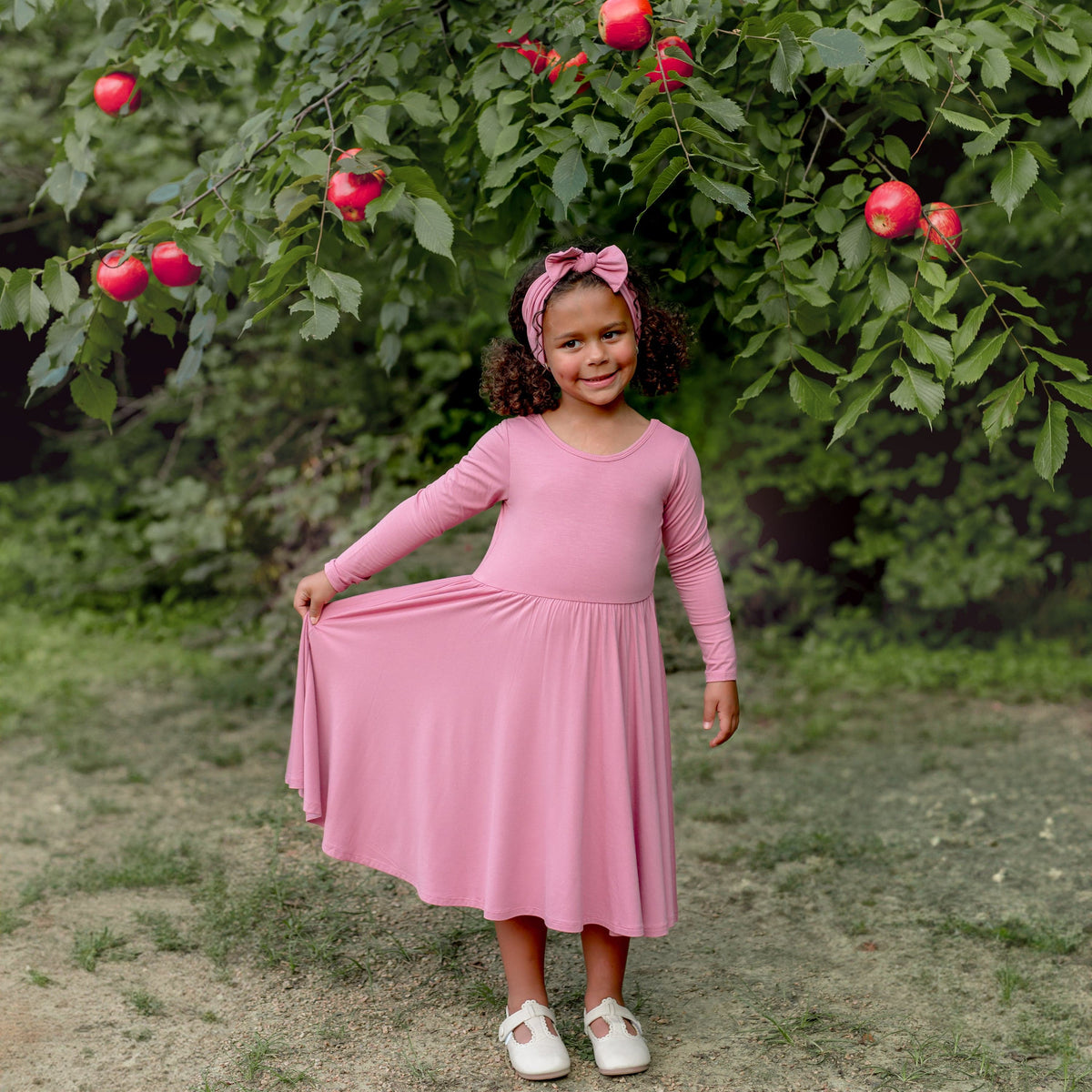 Young girl standing under an apple tree in the grass holding the the Long Sleeve Twirl Dress in Apple Blossom out with her hand