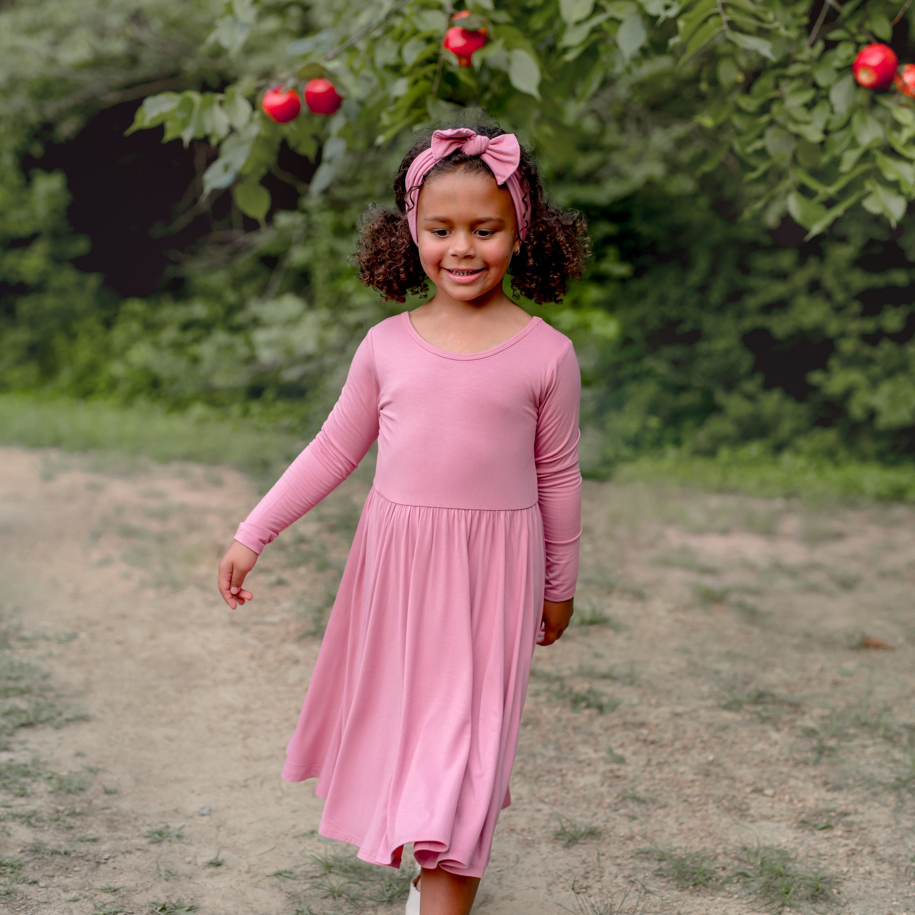 Young girl walking in a field in front of an apple tree wearing the Long Sleeve Twirl Dress in Apple Blossom and matching knotted bow headband