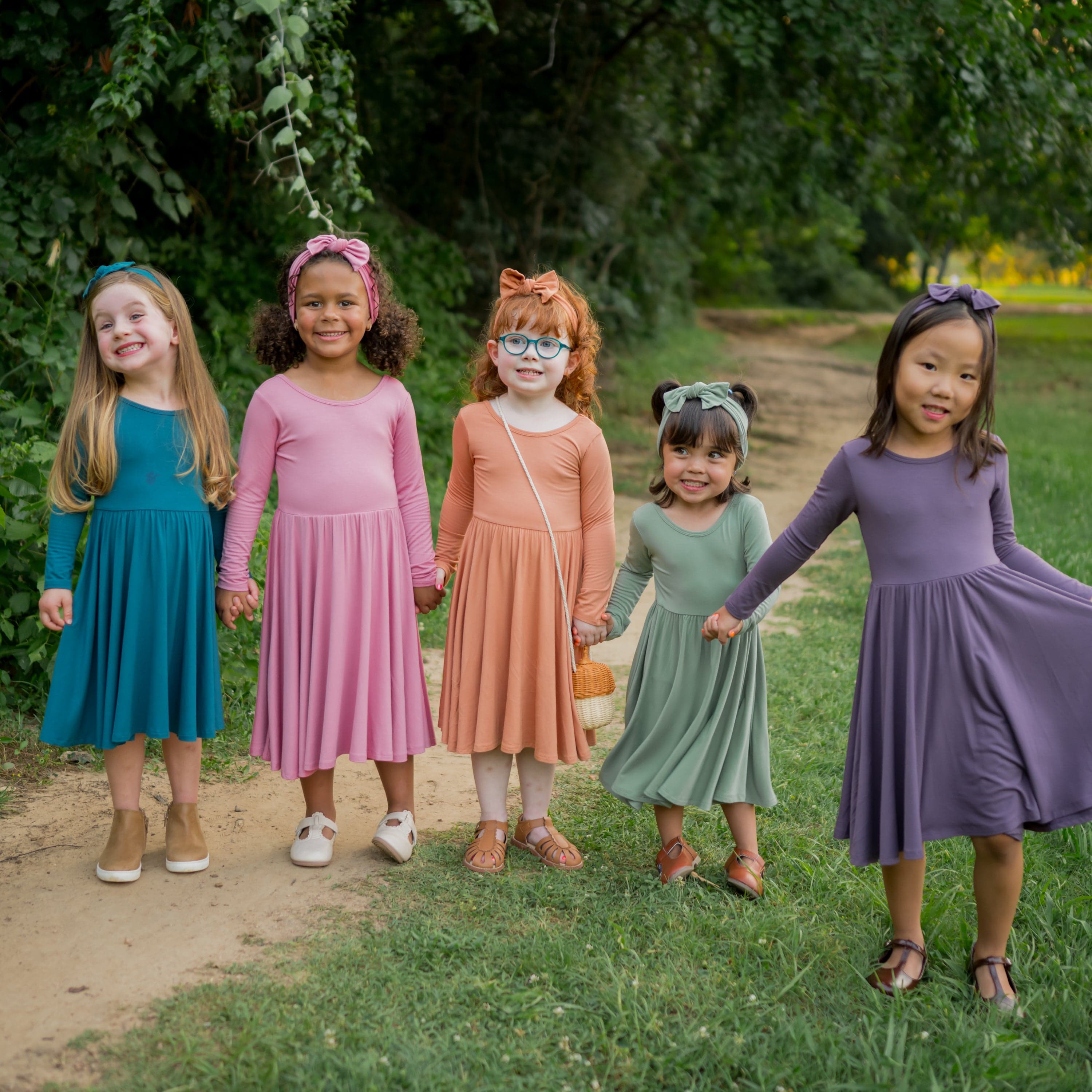 Five young girls standing outside in the grass holding hands wearing various colors in the long sleeve twirl dresses