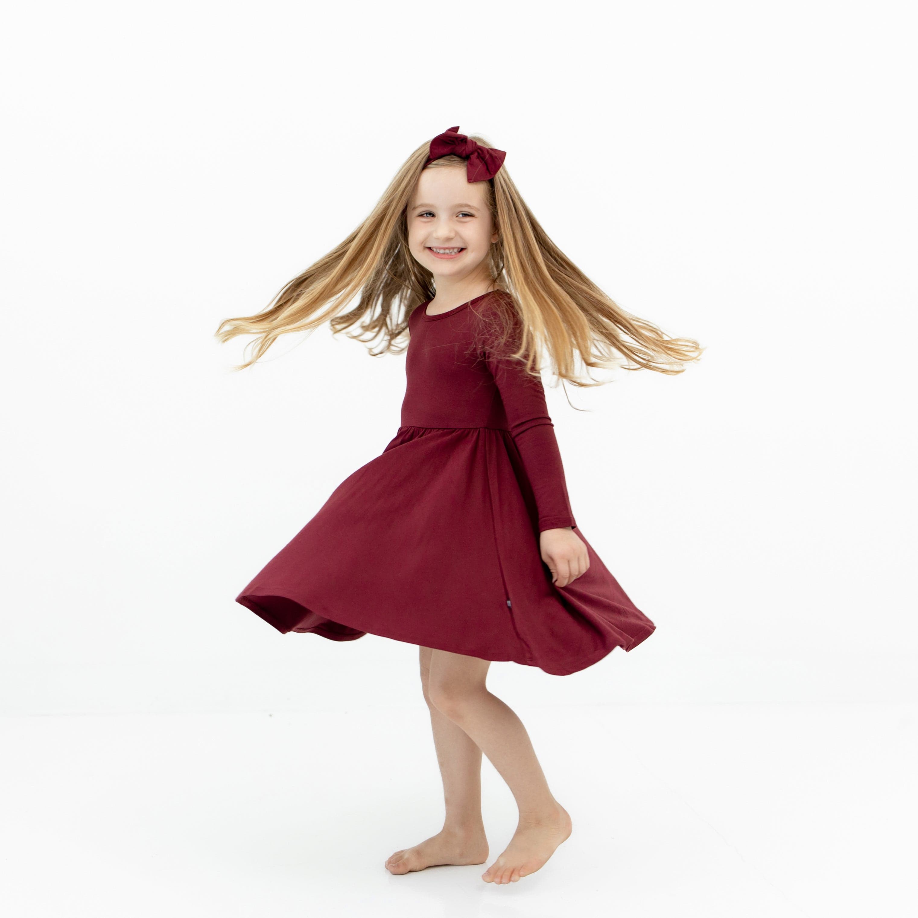 Smiling girl twirling in the Long Sleeve Twirl Dress in Burgundy wearing a matching knotted bow headband
