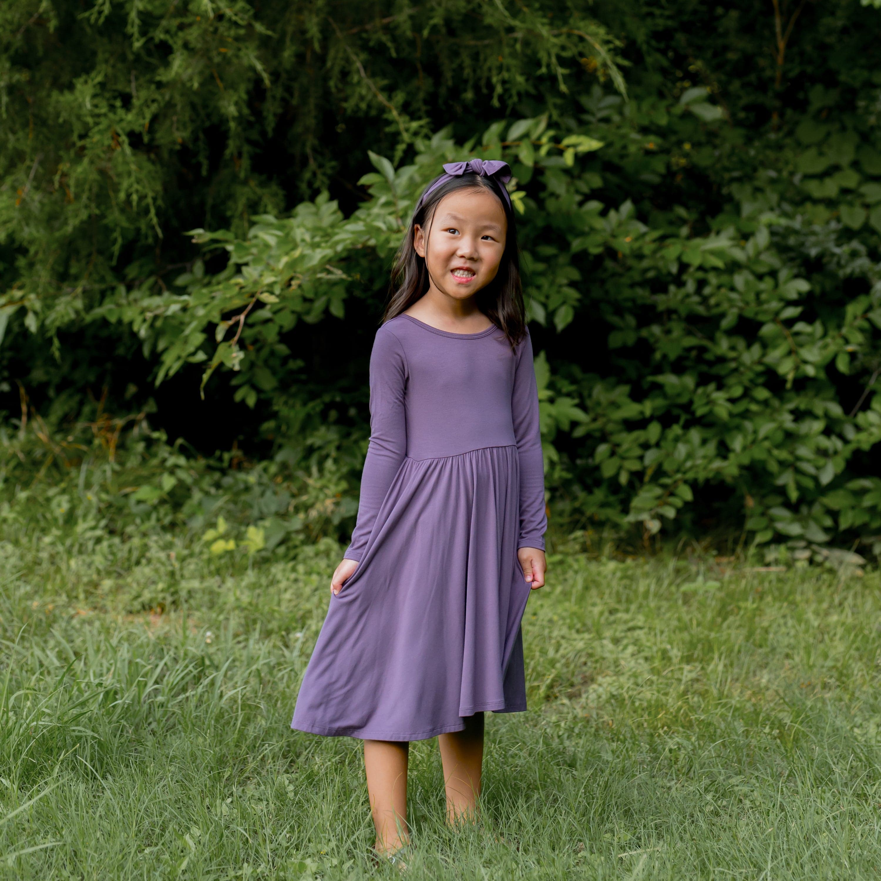 Young girl standing in the grass in front of trees wearing the Long Sleeve Twirl Dress in Currant with matching knotted bow headband