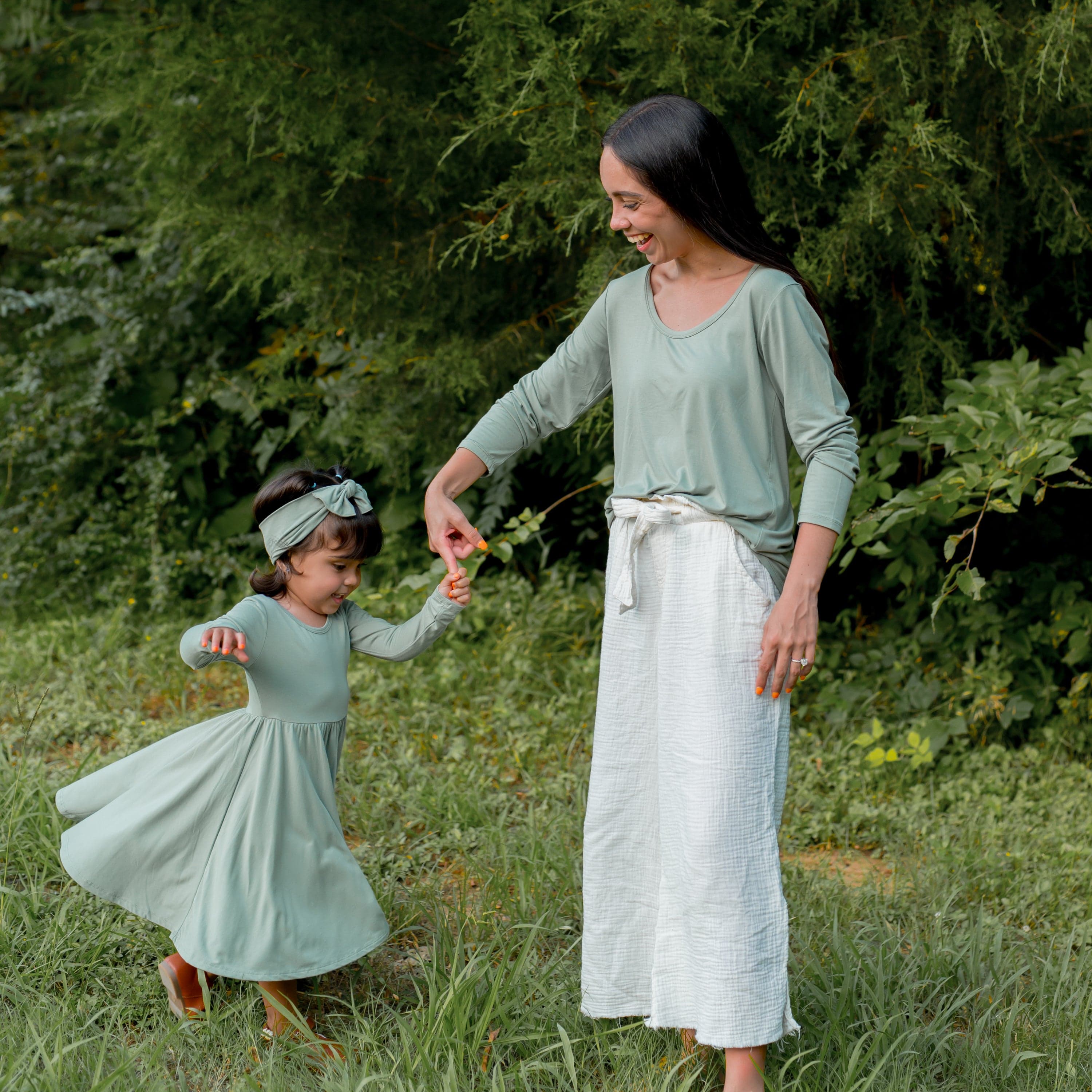 Young girl wearing the Long Sleeve Twirl Dress in Thyme being twirled by her mother standing in a grass field with trees behind them