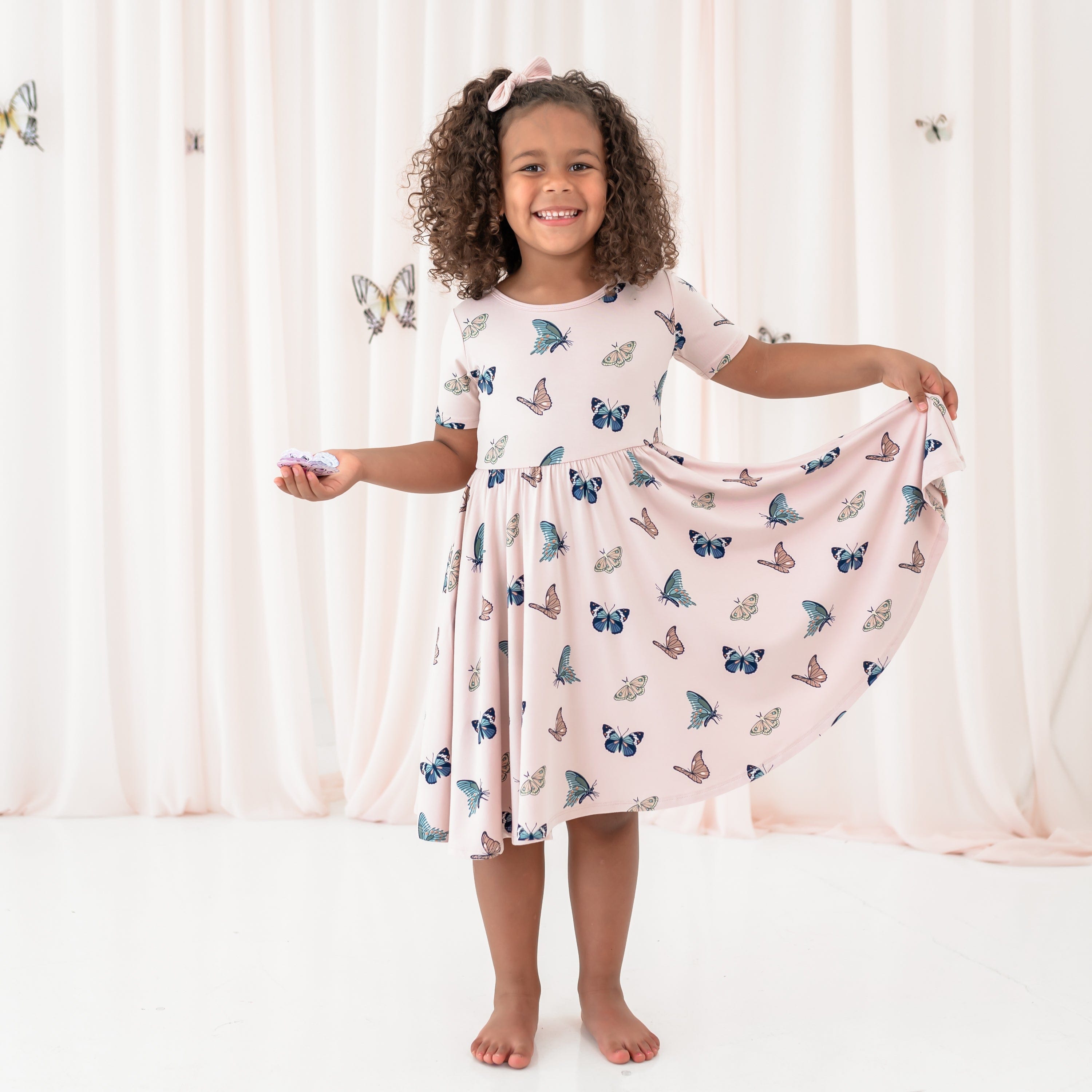 Young girl wearing the Twirl Dress in Blush Butterfly holding the bottom hem of the twirl skirt out at her side