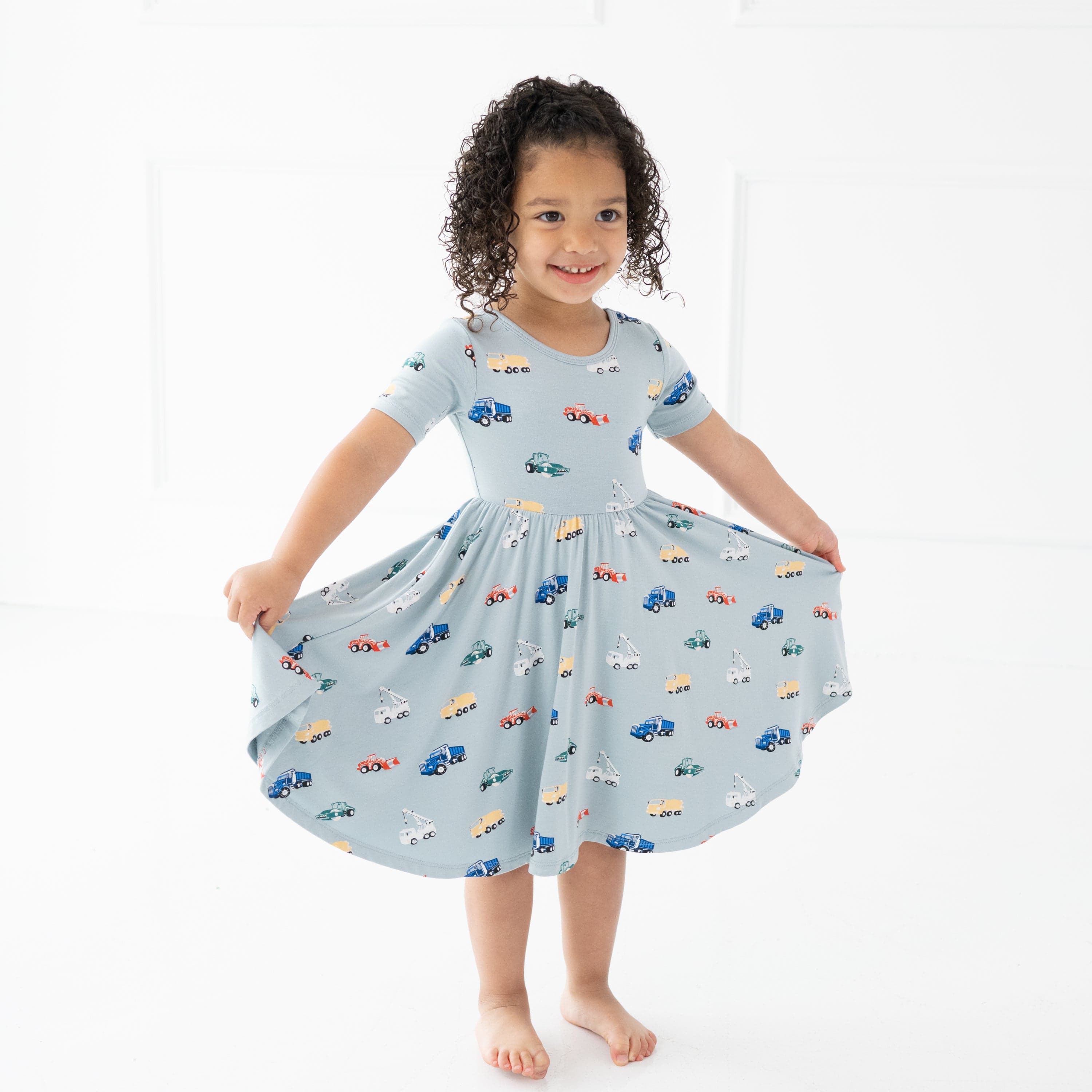 Toddler girl standing wearing the Twirl Dress in Construction holding the bottom hem of the twirl skirt out with both her hands
