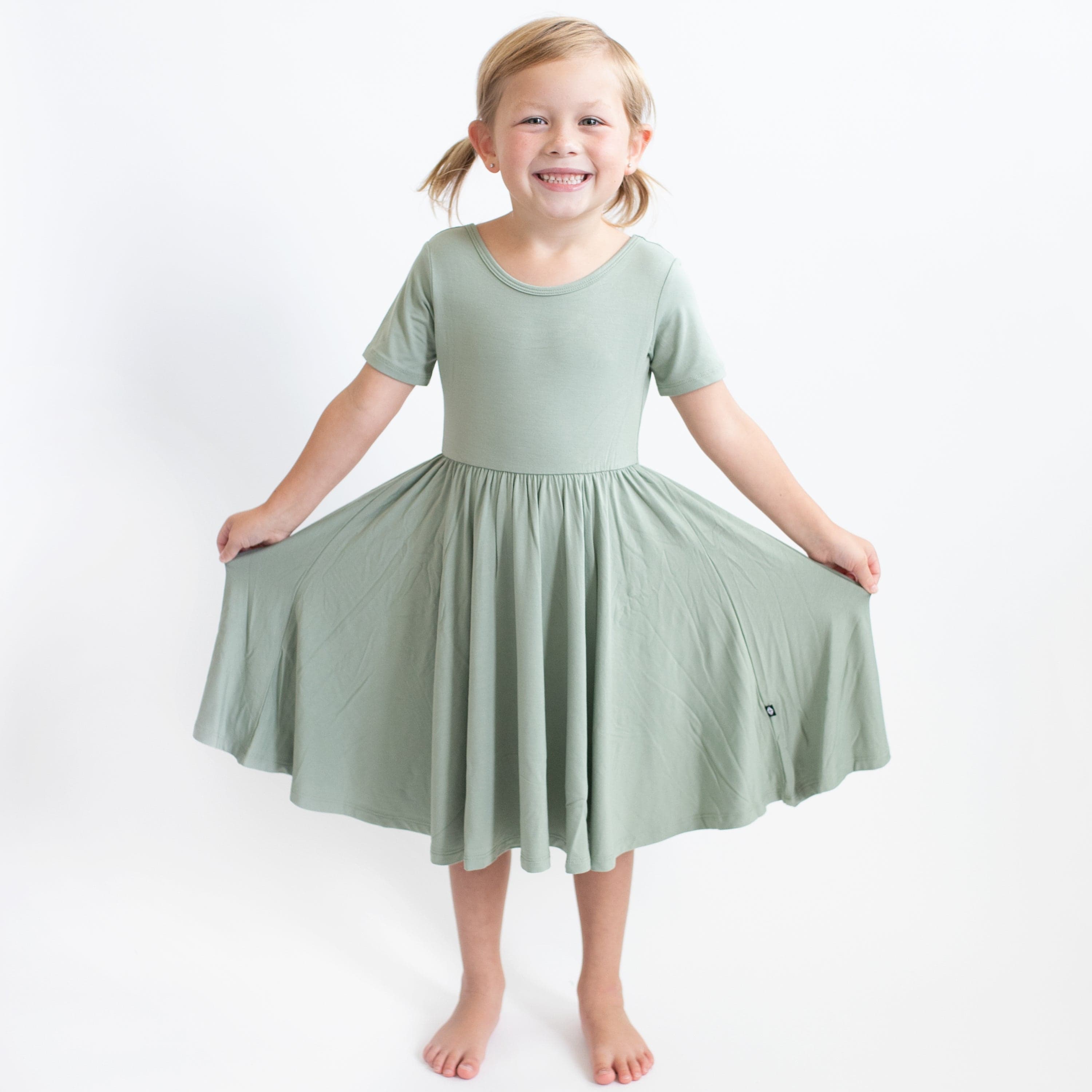 Young girl modeling the Twirl Dress in Thyme holding out the twirl skirt with both hands