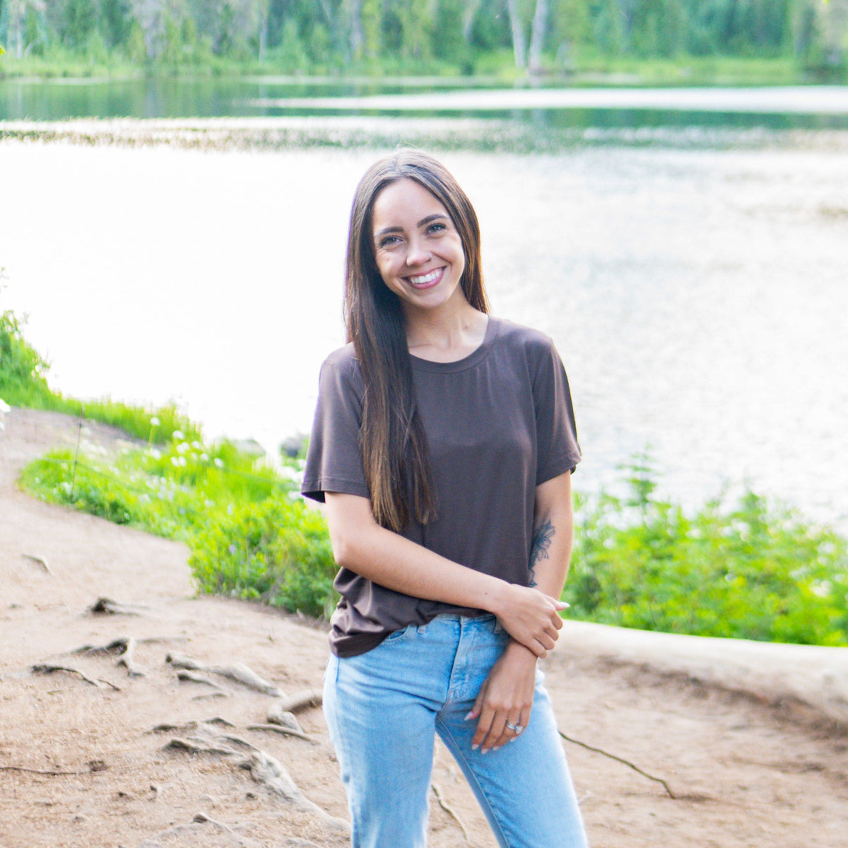 Smiling female model standing on a path near the waters edge wearing the Women's Crew Neck Tee in Espresso paired with jeans