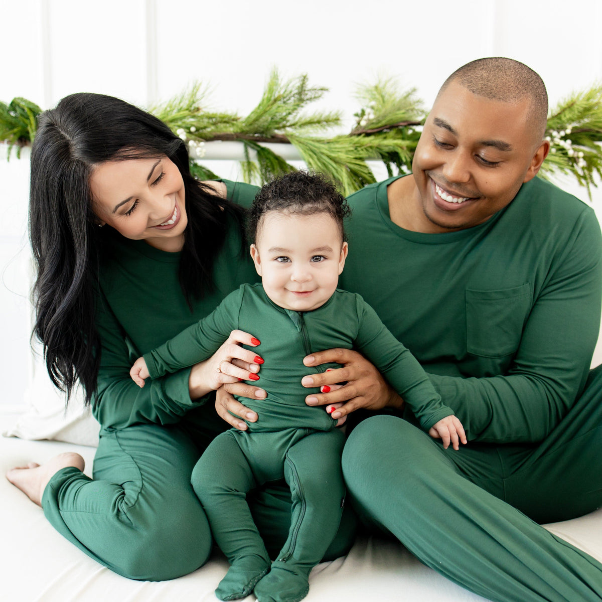 Mom and Dad wearing Evergreen Jogger Sets sitting side by side holding baby between them who is wearing the Zippered Footie in Evergreen