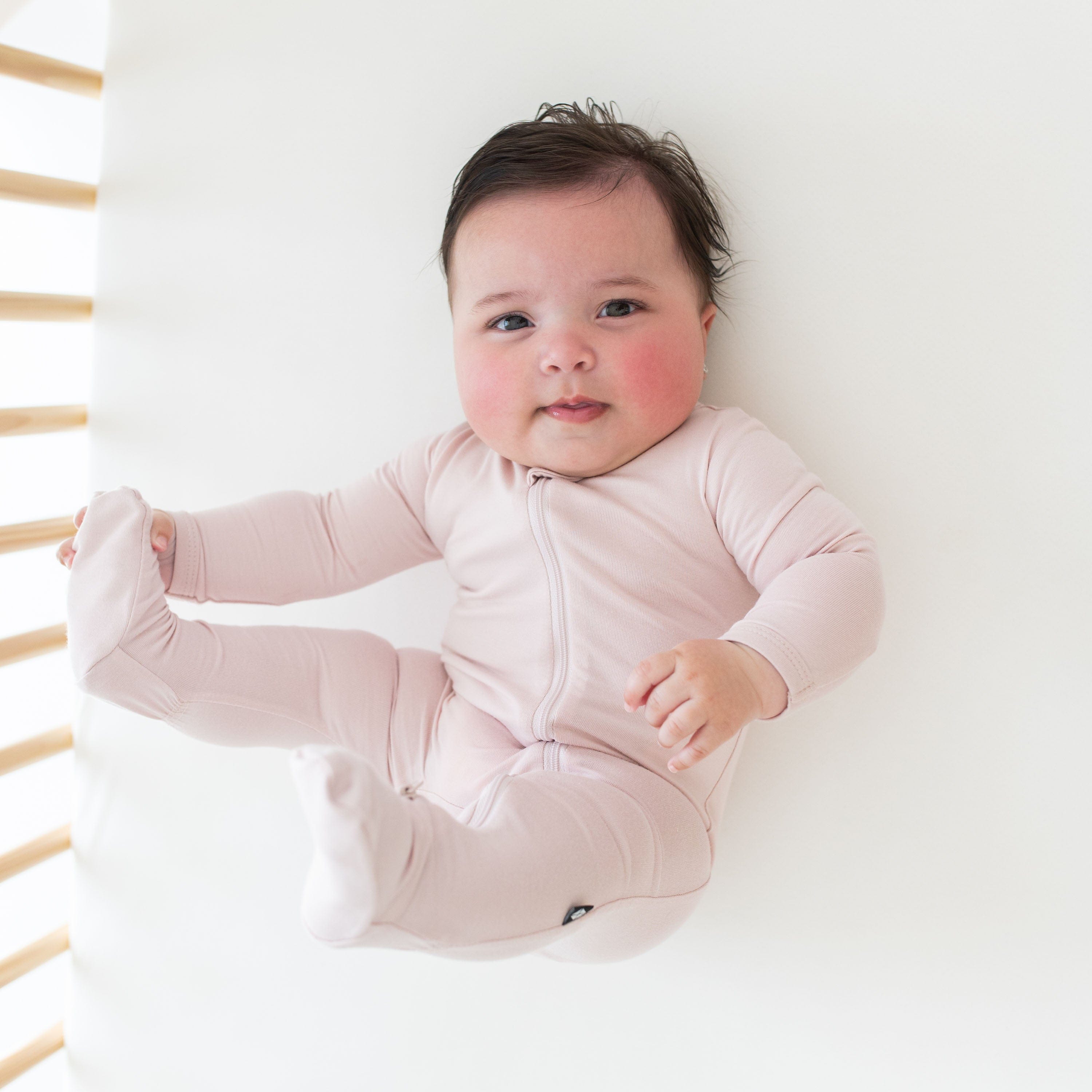 Baby holding its foot while laying on a crib sheet in Oat and a Zippered Footie in Blush
