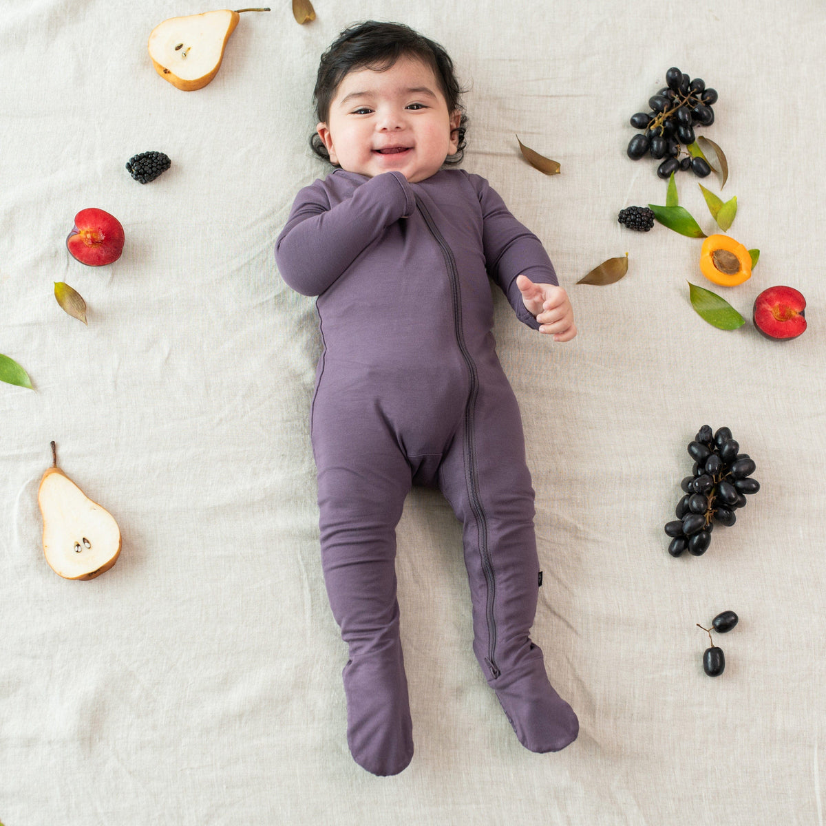 Infant wearing the Zippered Footie in Currant surrounded by real fruit