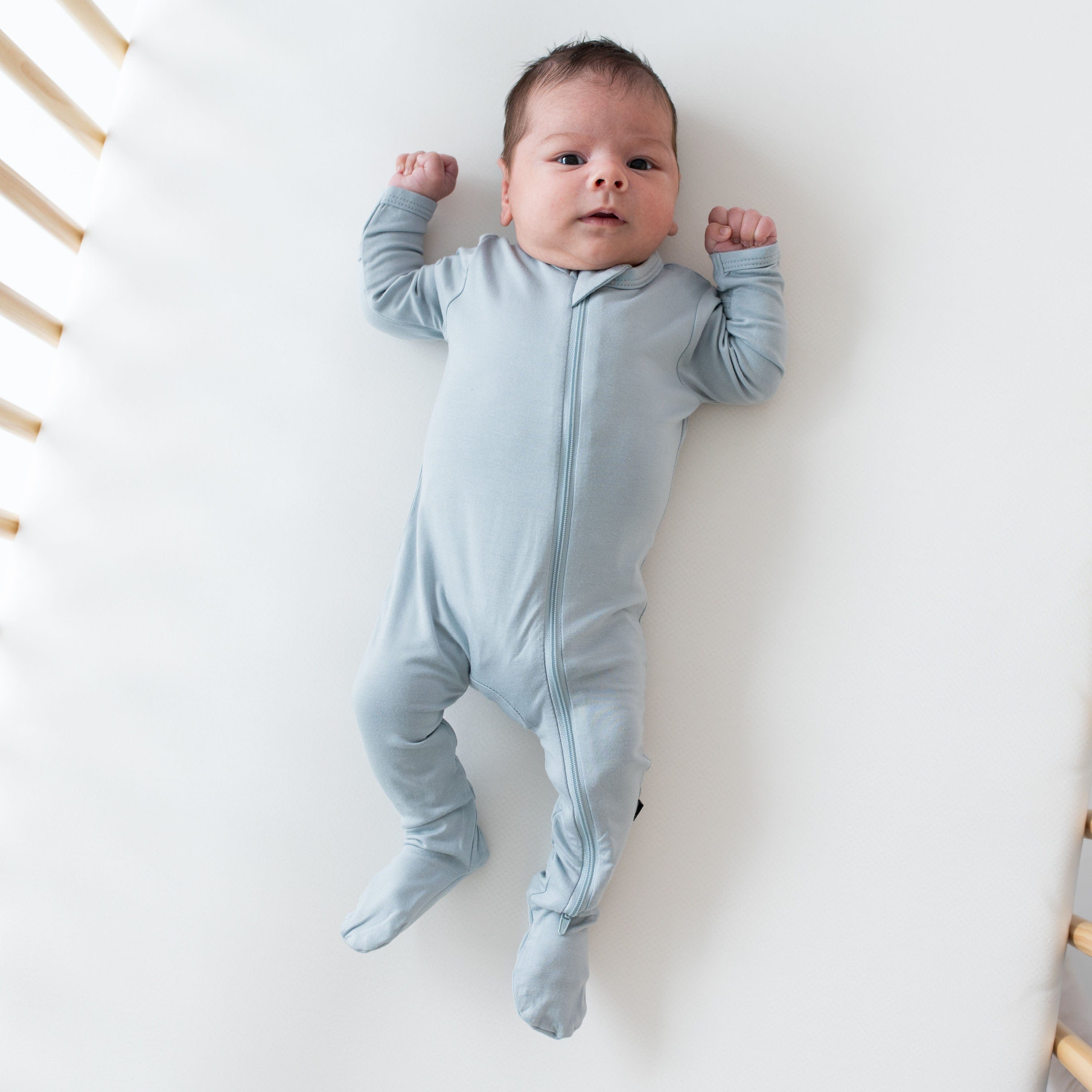 Newborn laying in a crib while wearing a Zippered Footie in Fog