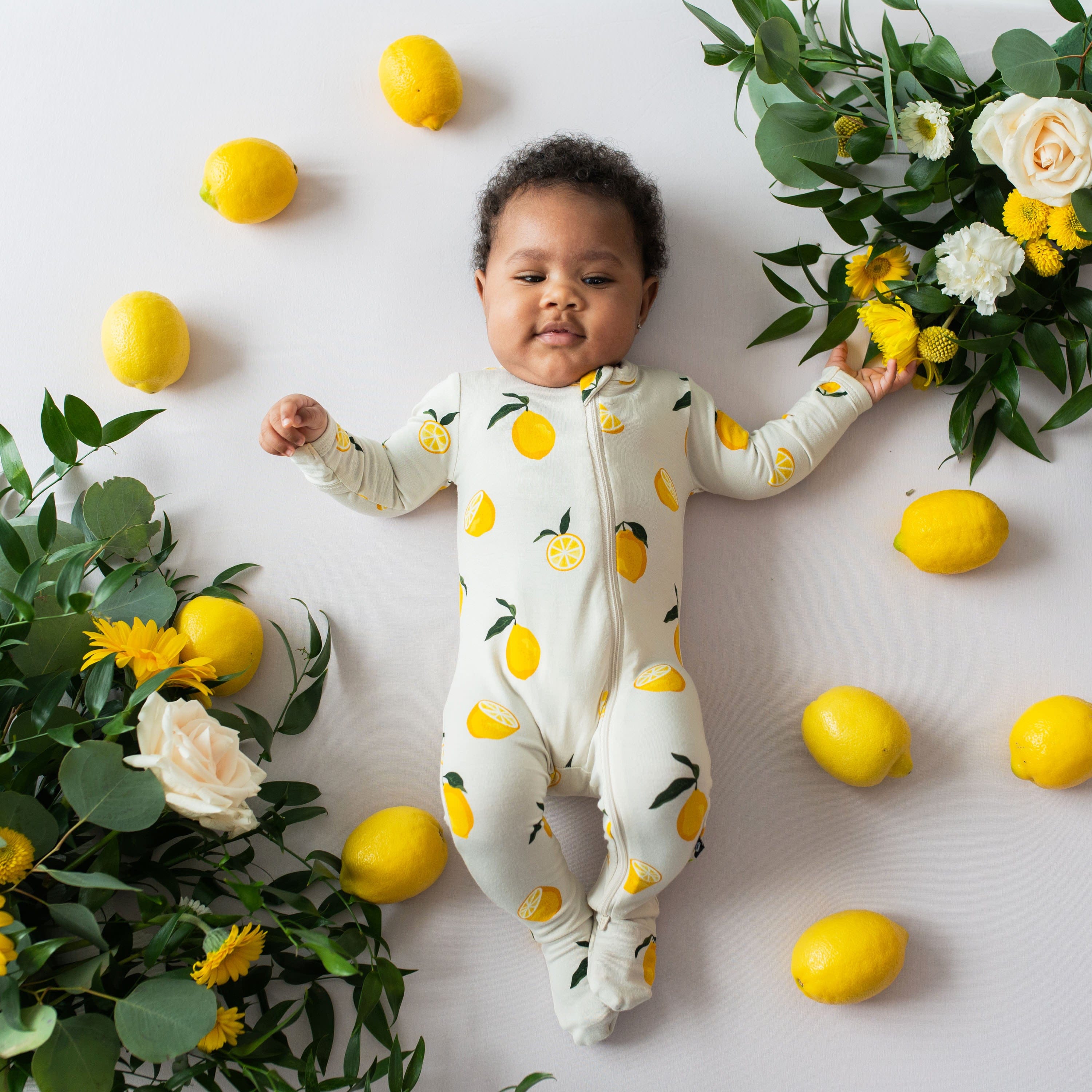 Infant wearing the Zippered Footie in Lemon surrounded by lemons and flowers