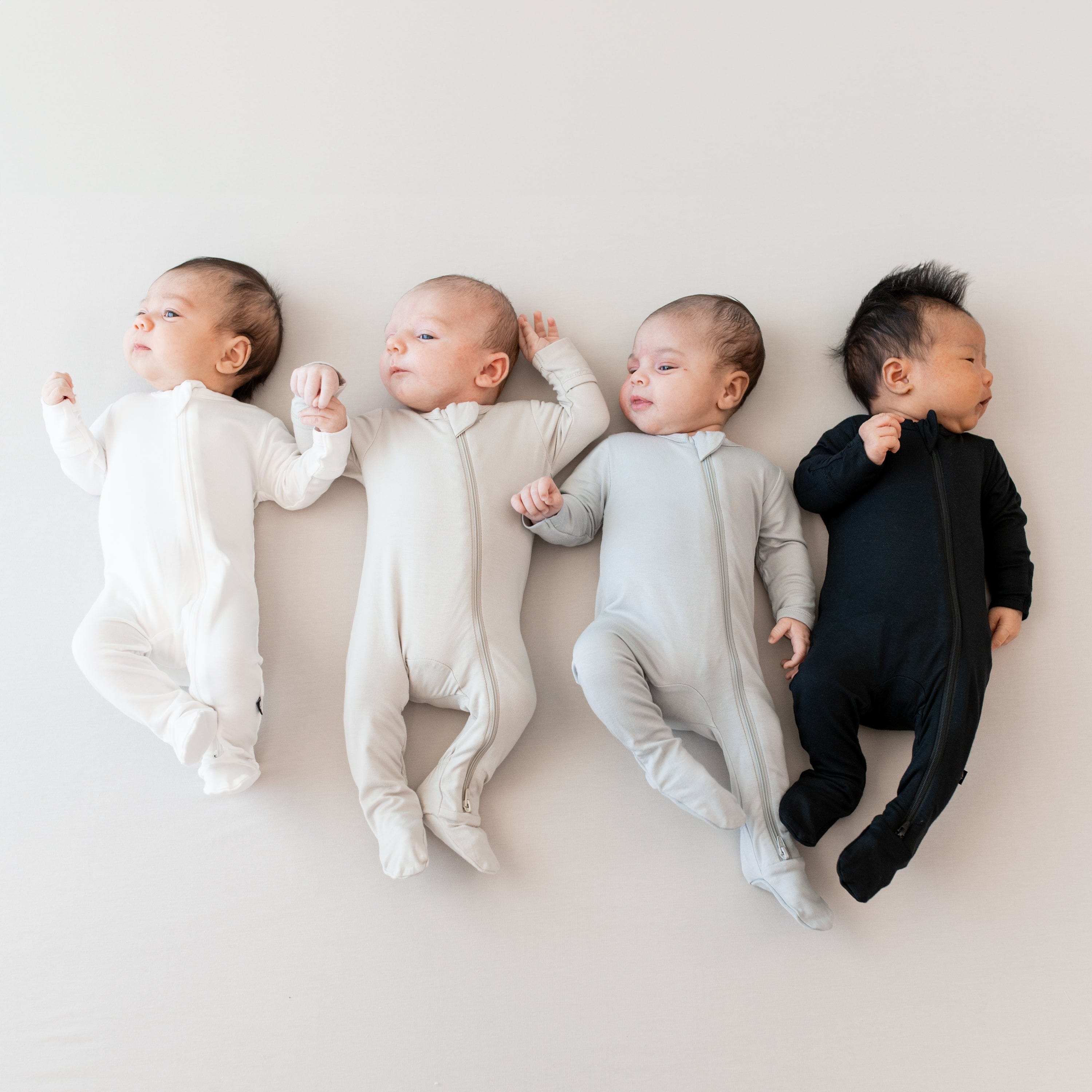 Four infants laying next to each other wearing Zippered Footies in Cloud, Oat, Storm and Midnight