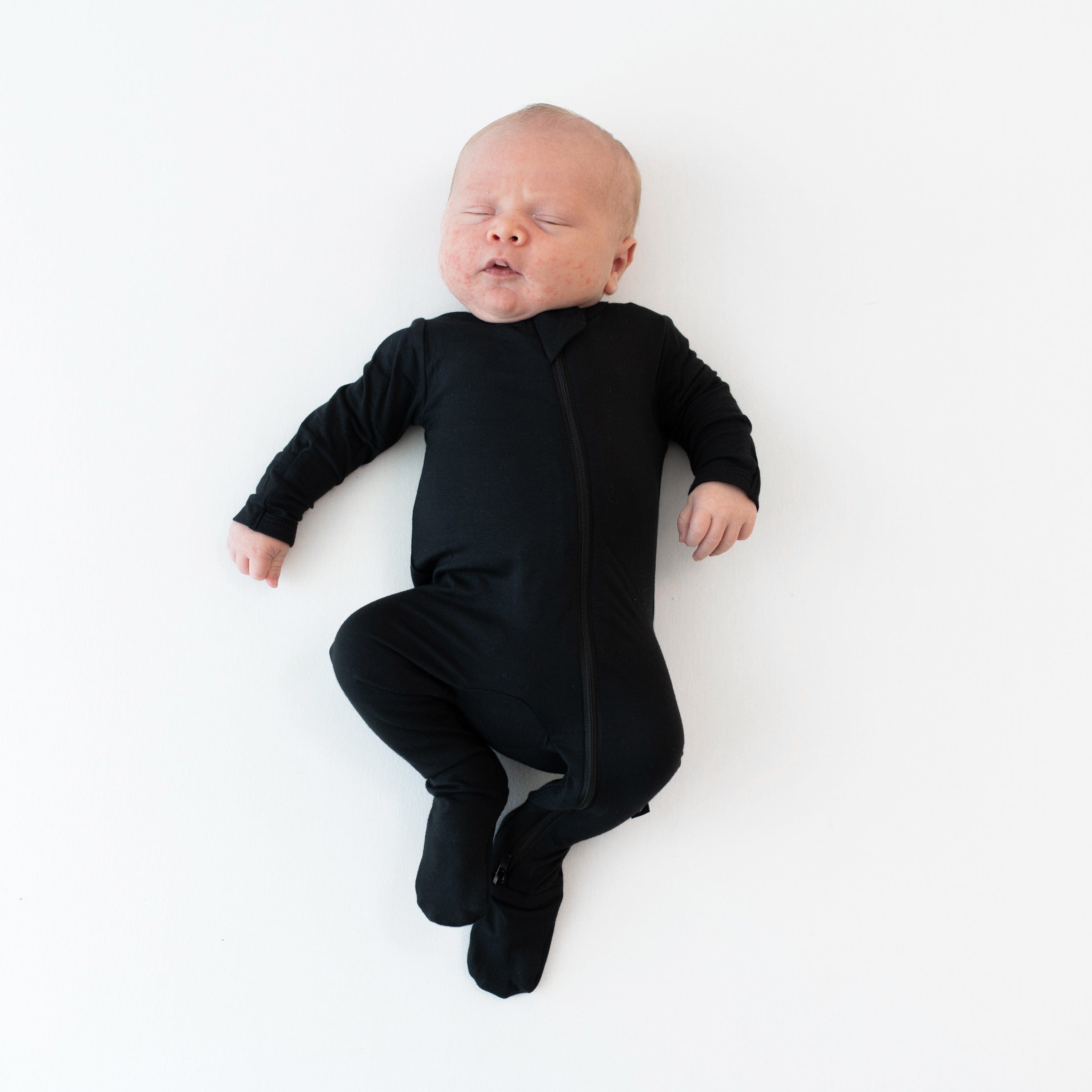 Sleeping infant wearing the Zippered Footie in Midnight while laying on a white surface
