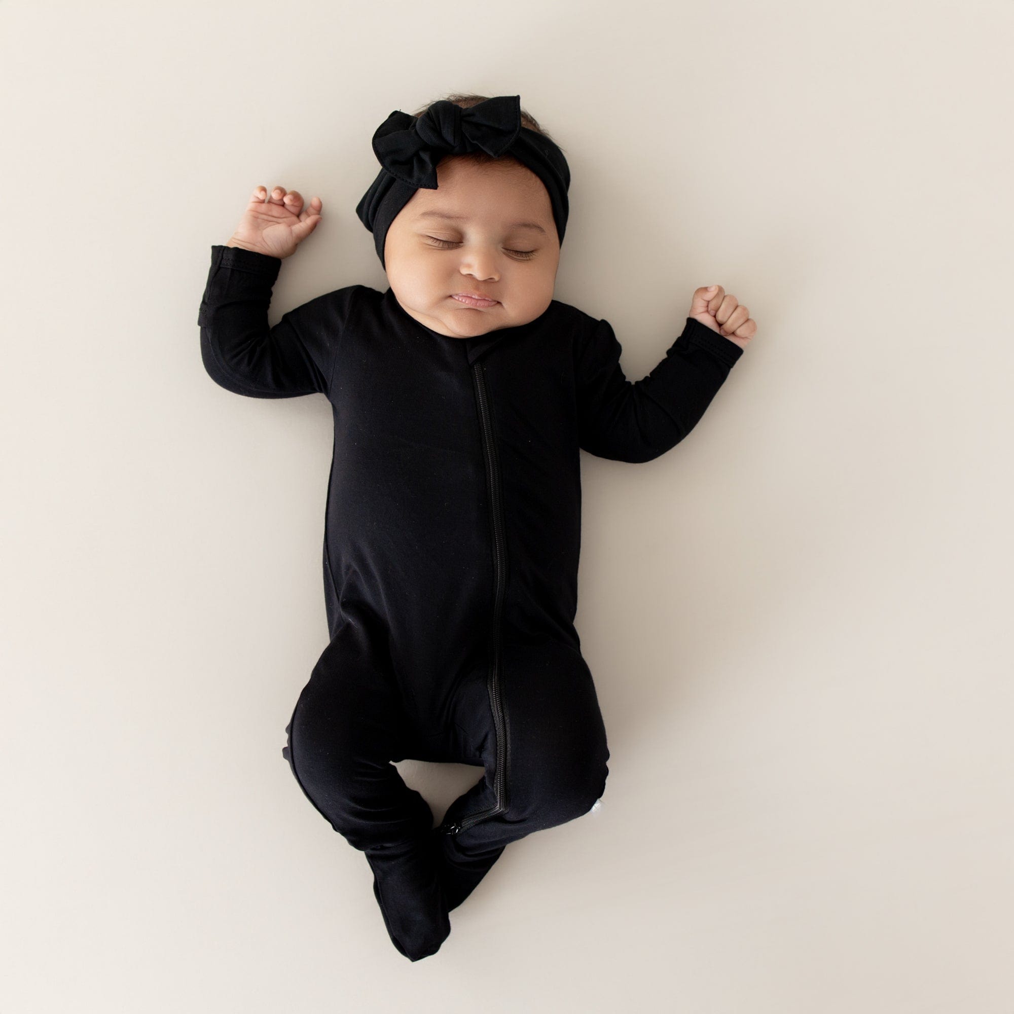 Infant model sleeping wearing the Zippered Footie in Midnight with the matching knotted bow