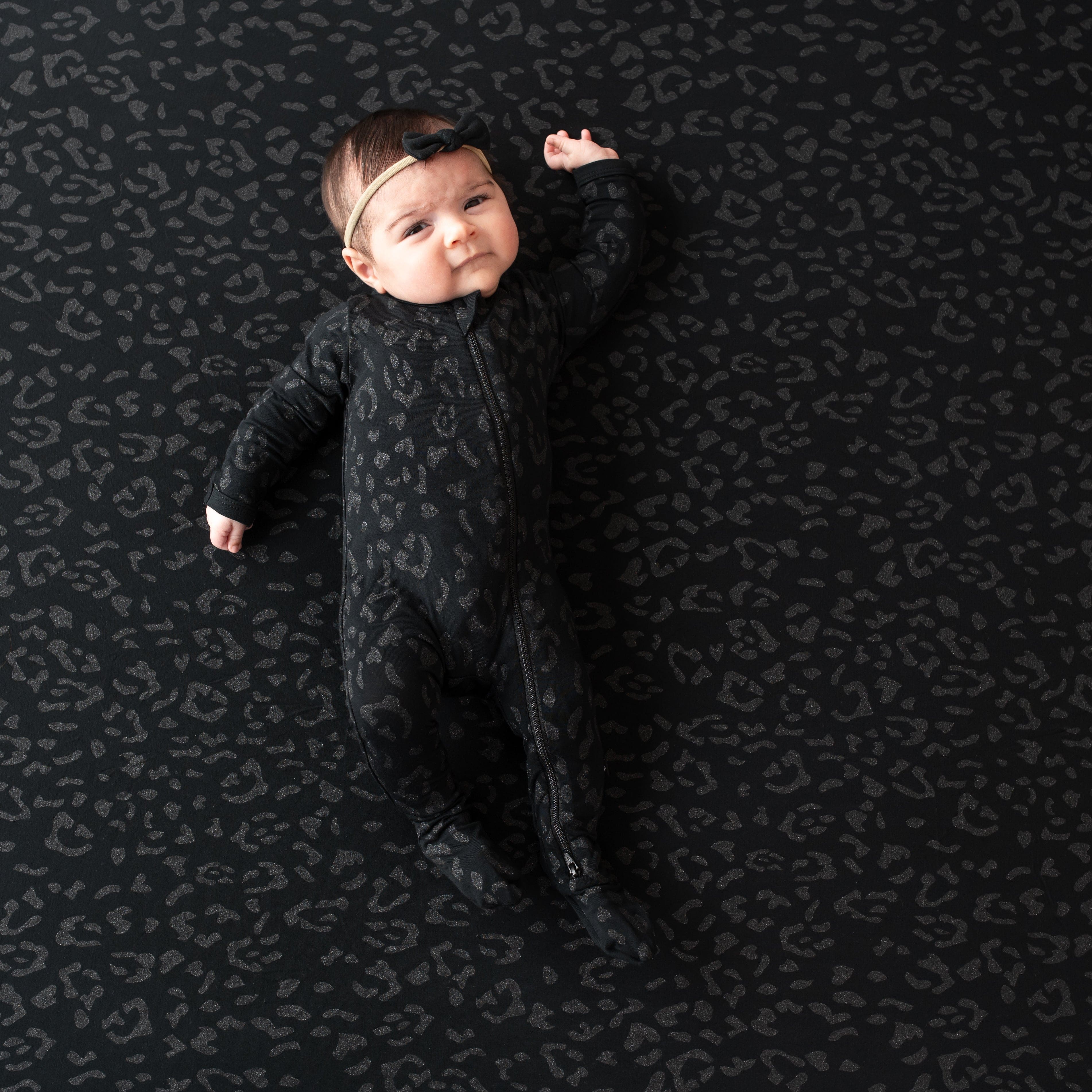 Infant modeling the Zippered Footie in Midnight Leopard with one hand up laying on a matching blanket