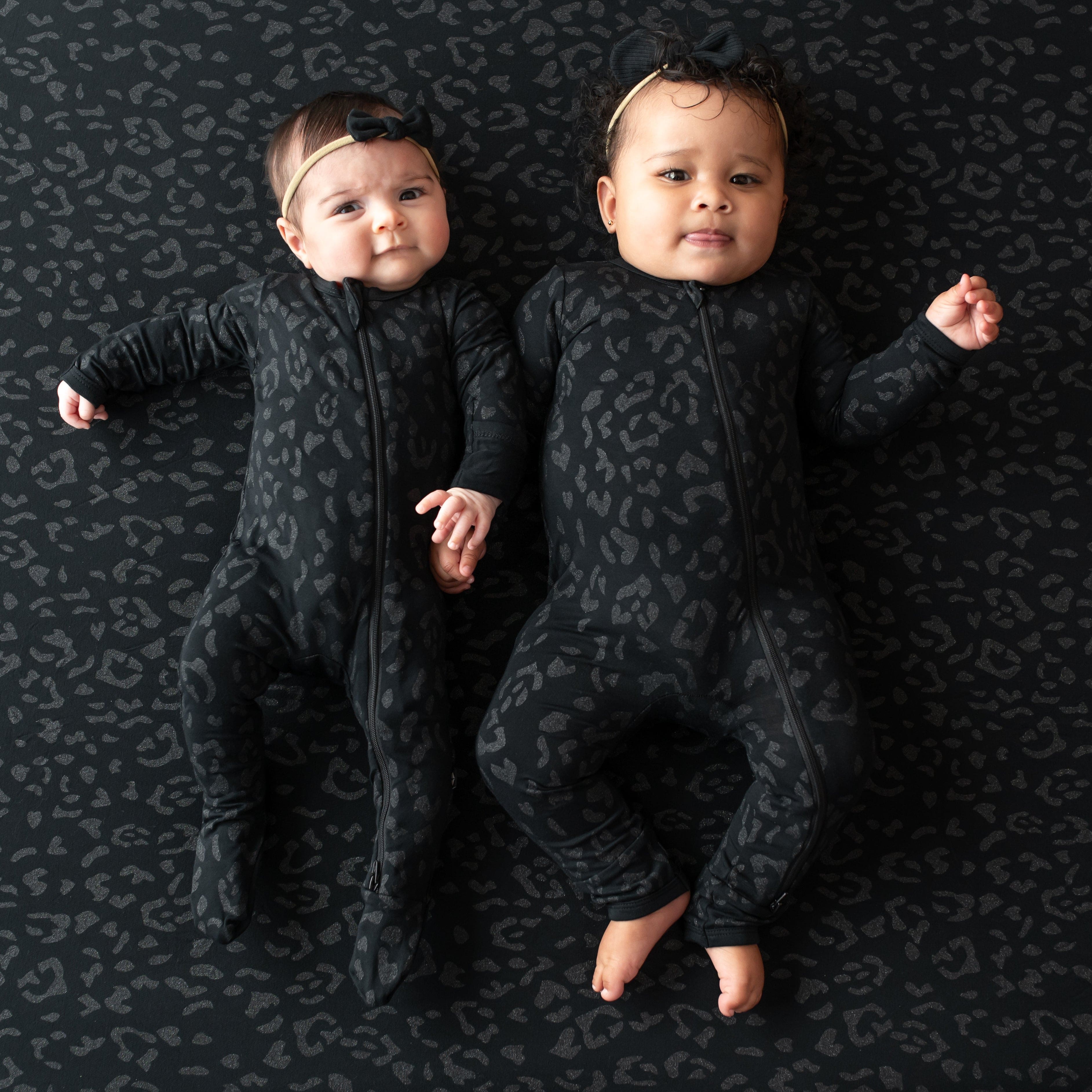 Two infant models wearing a zippered footie and zippered romper in Midnight Leopard