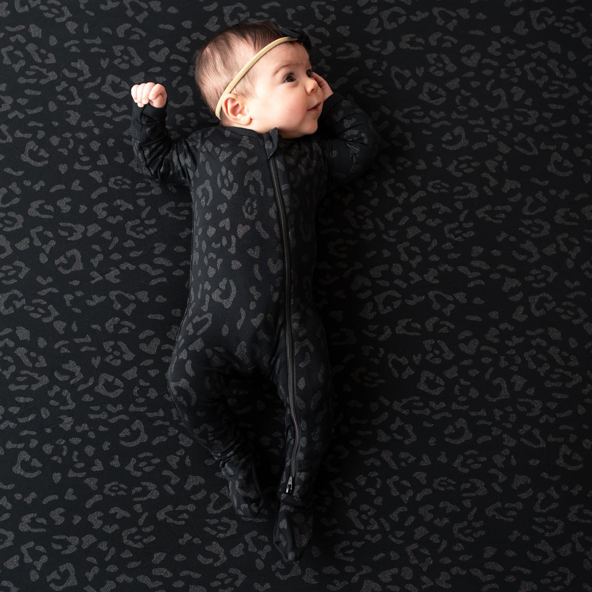 Infant baby looking off to the side wearing the Zippered Footie in Midnight Leopard laying on a matching blanket