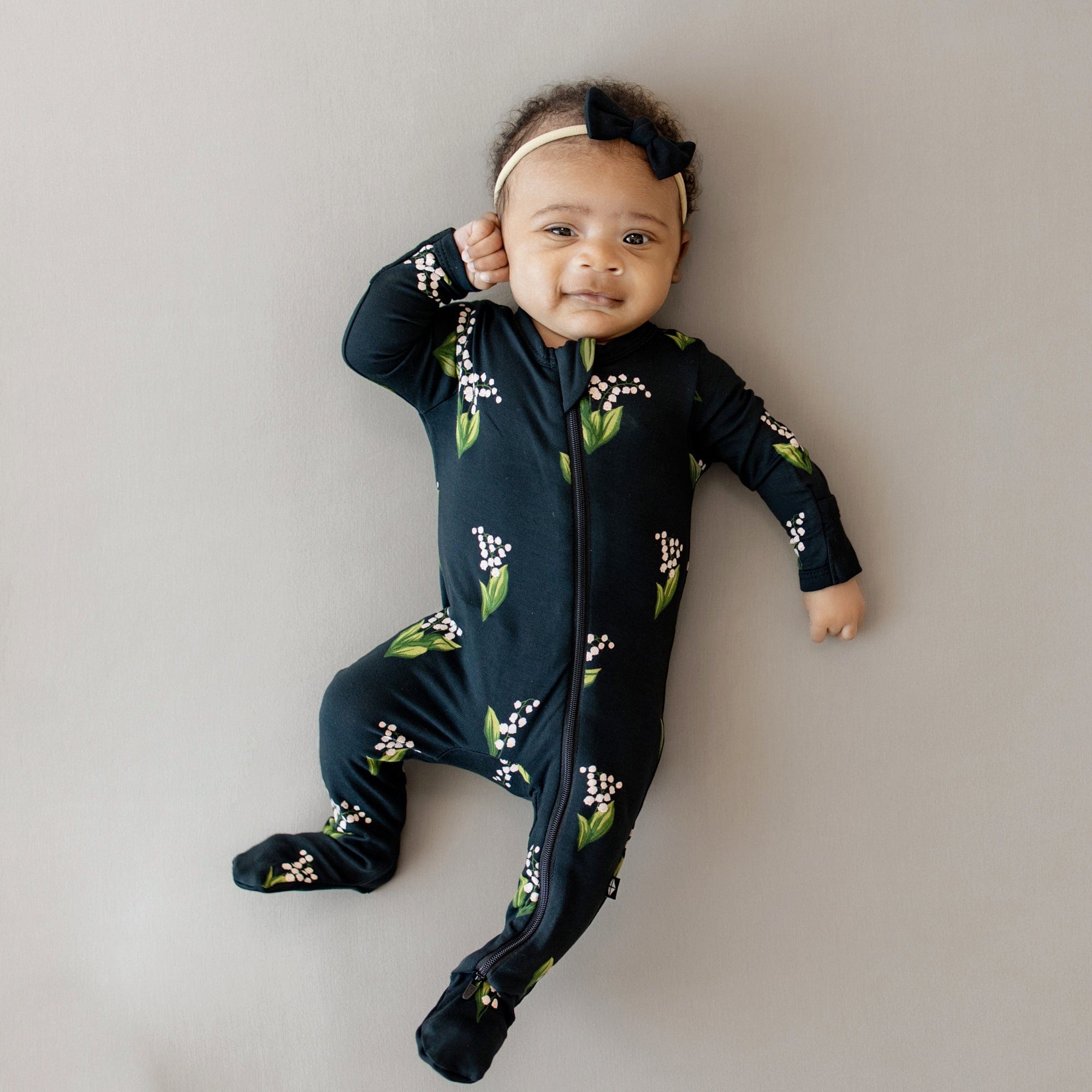 Infant wearing the Zippered Footie in Midnight Lily with one hand up to the side of her face