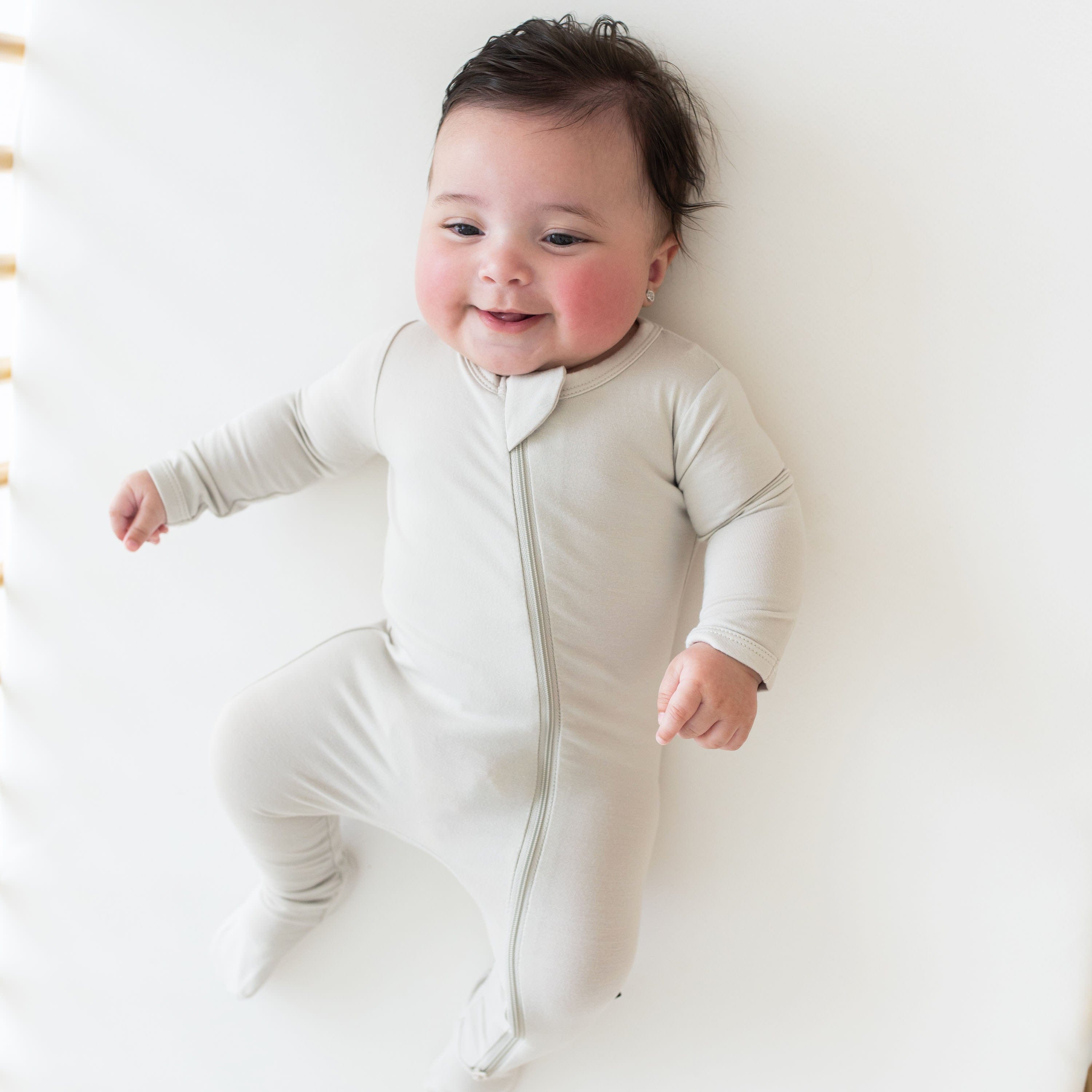 Smiling baby wearing a Zippered Romper in Oat and laying on matching surface