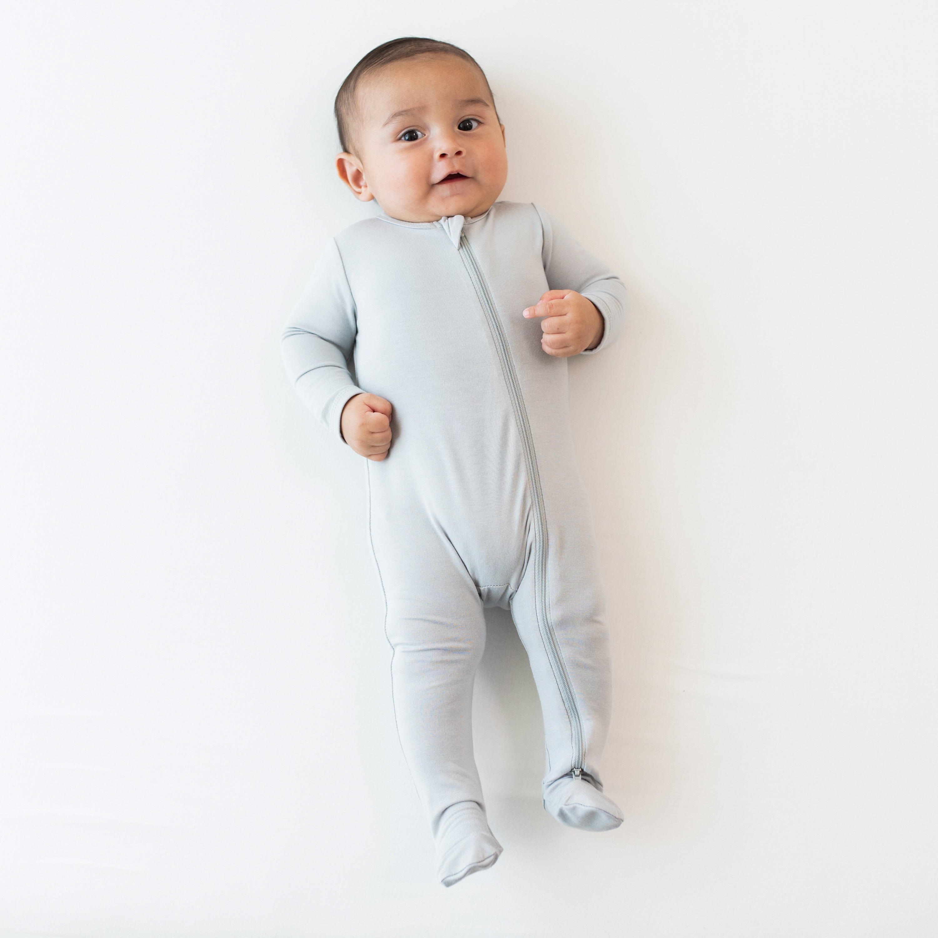 Infant laying on an off-white surface while wearing a Zippered Footie in Storm