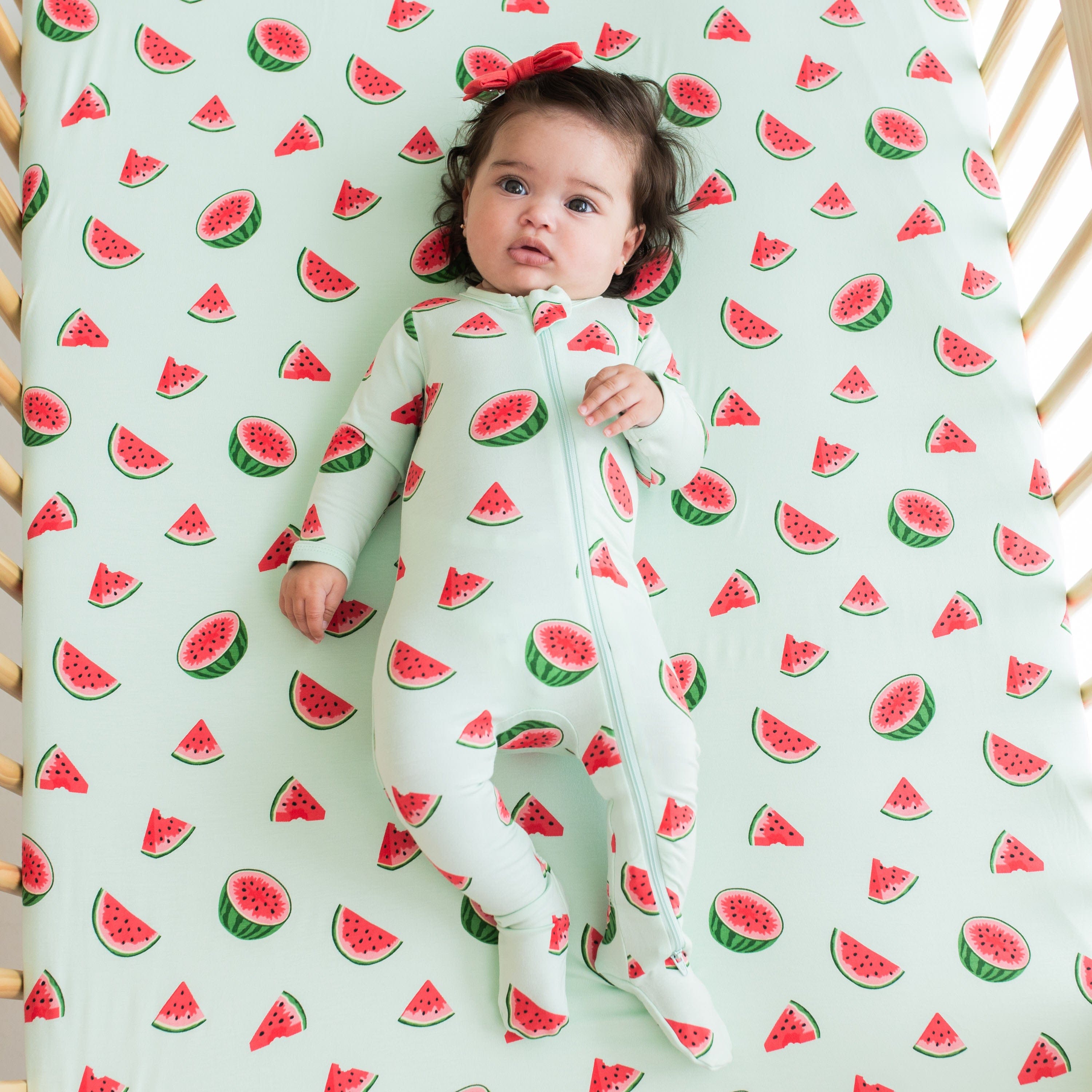 Infant wearing the Zippered Footie in Watermelon laying on matching crib sheet