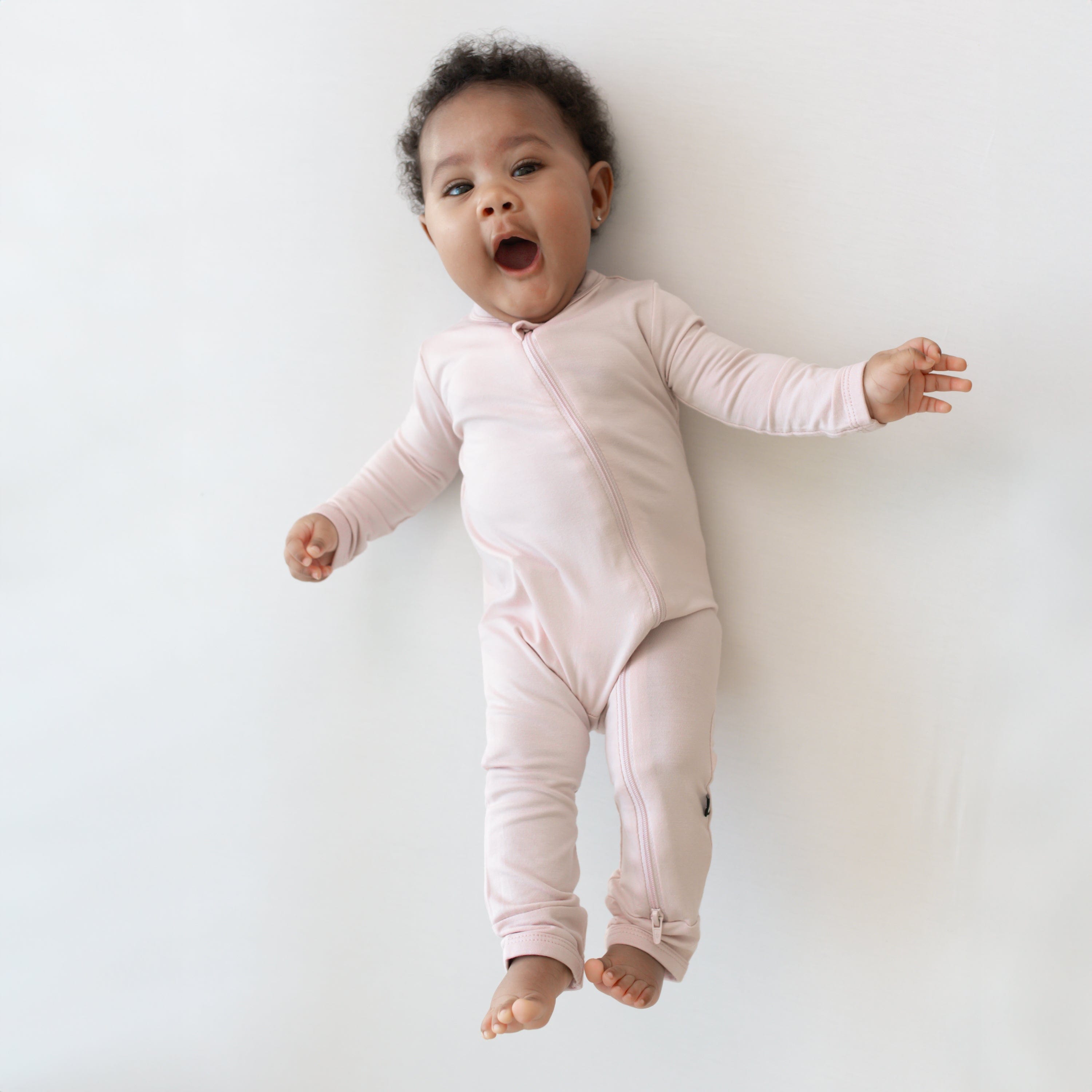 Infant laying on an off-white surface while wearing a Zippered Romper in Blush