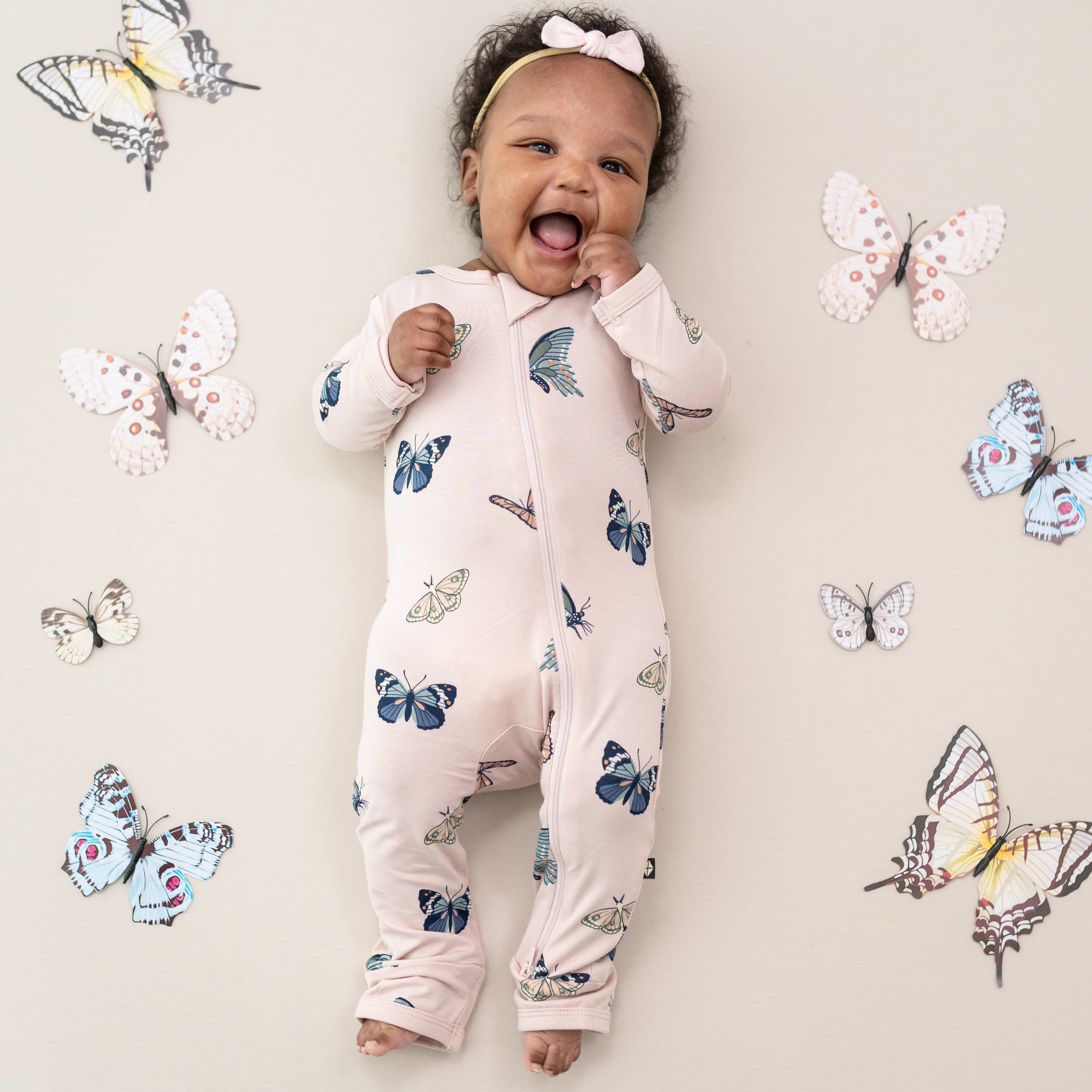 Smiling baby wearing the Zippered Romper in Blush Butterfly surrounded by toy butterflies