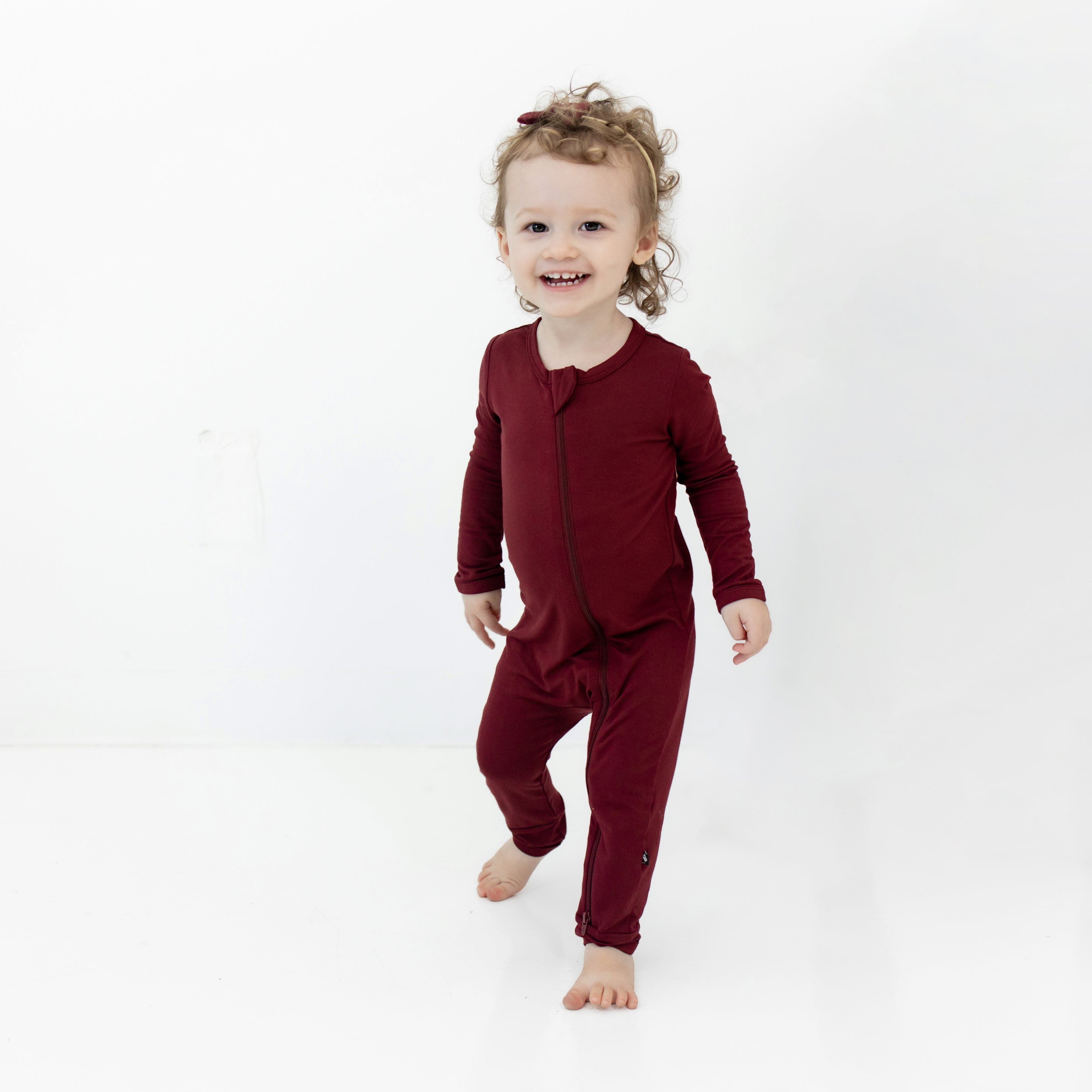 Toddler standing wearing the Zippered Romper in Burgundy with matching tiny nylon bow