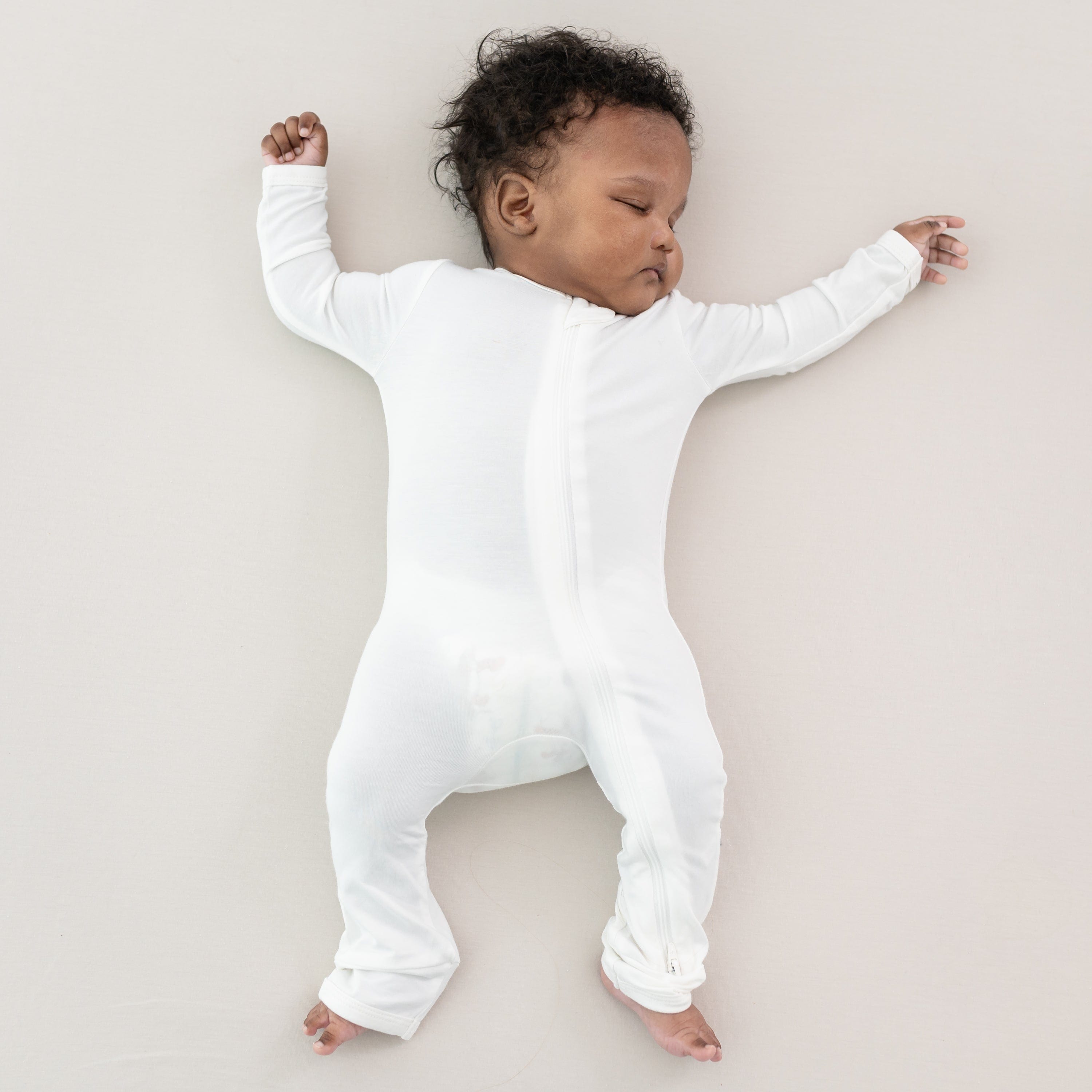 Sleeping baby laying on an Oat surface while wearing a Kyte Baby Zippered Romper in Cloud